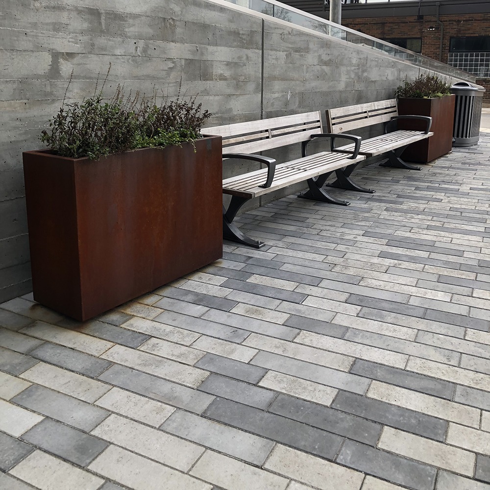 Concrete pathway with metal planters and wooden benches. Modern outdoor seating area near a building, featuring gray stone paving and minimalistic design.