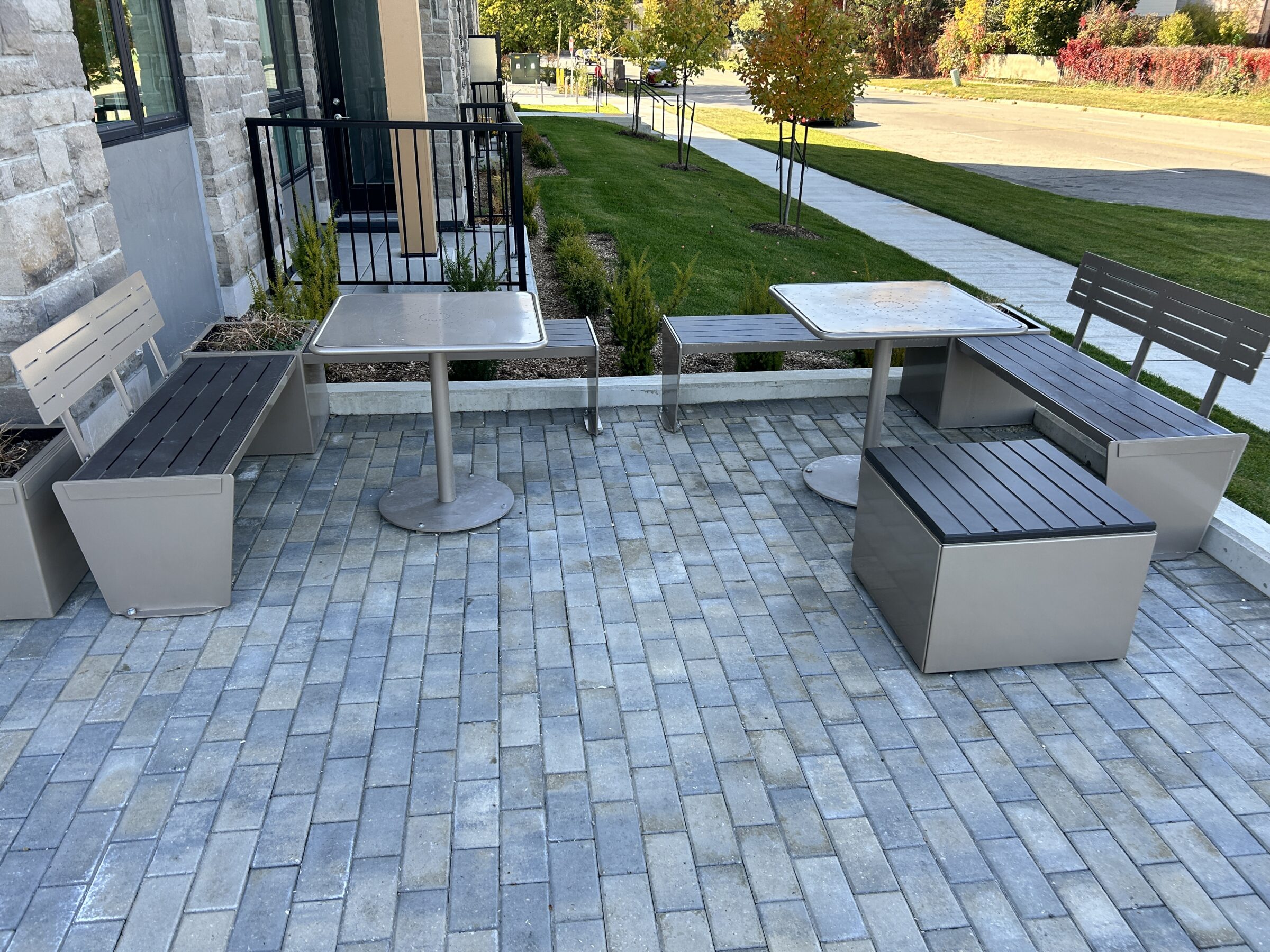 Modern outdoor seating area with benches and tables on a paved surface, surrounded by greenery and a sidewalk in an urban setting.