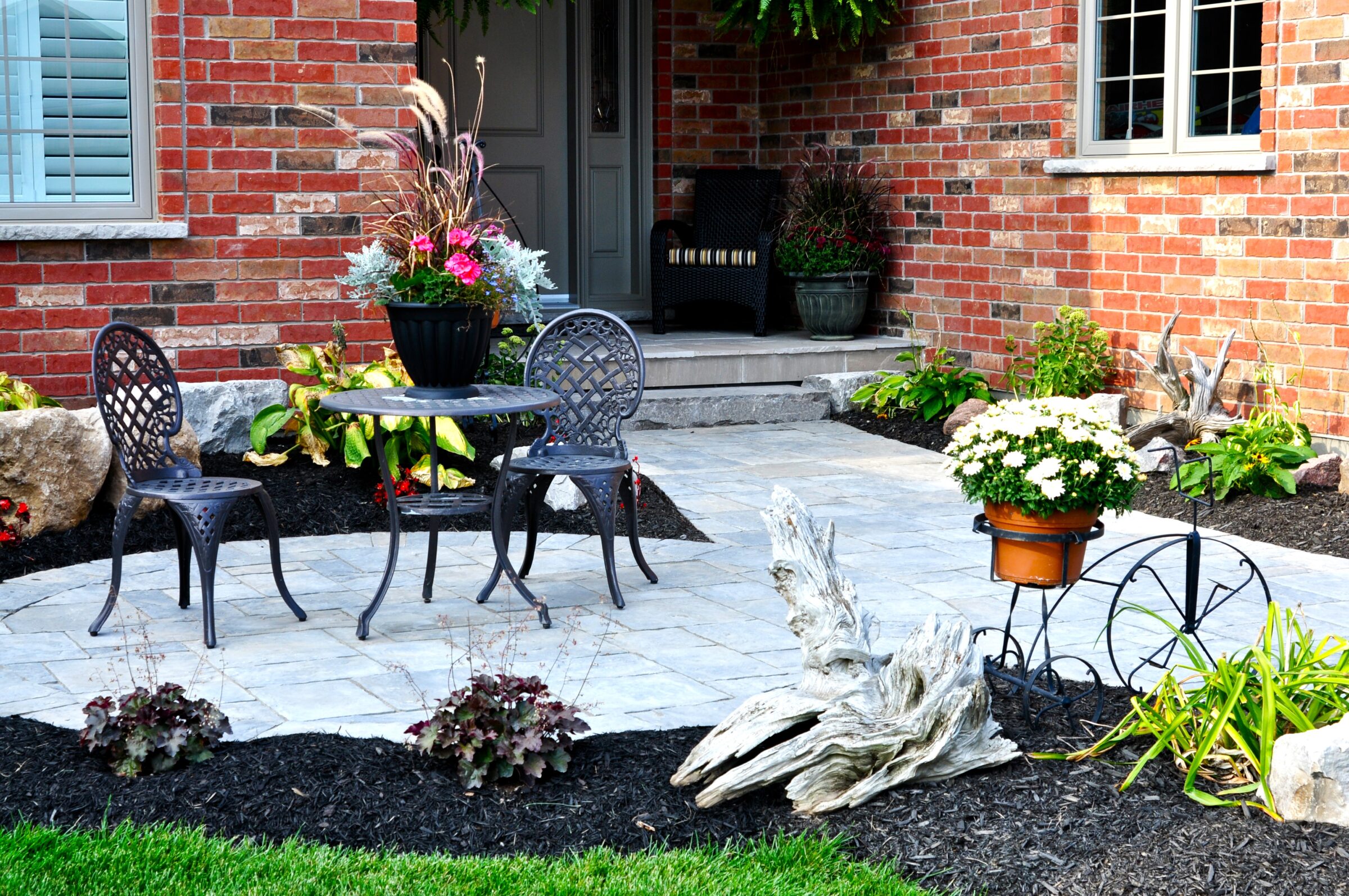 Brick house entrance with a patio set, decorative plants, and a bicycle planter. Neatly landscaped, featuring colorful flowers and greenery.