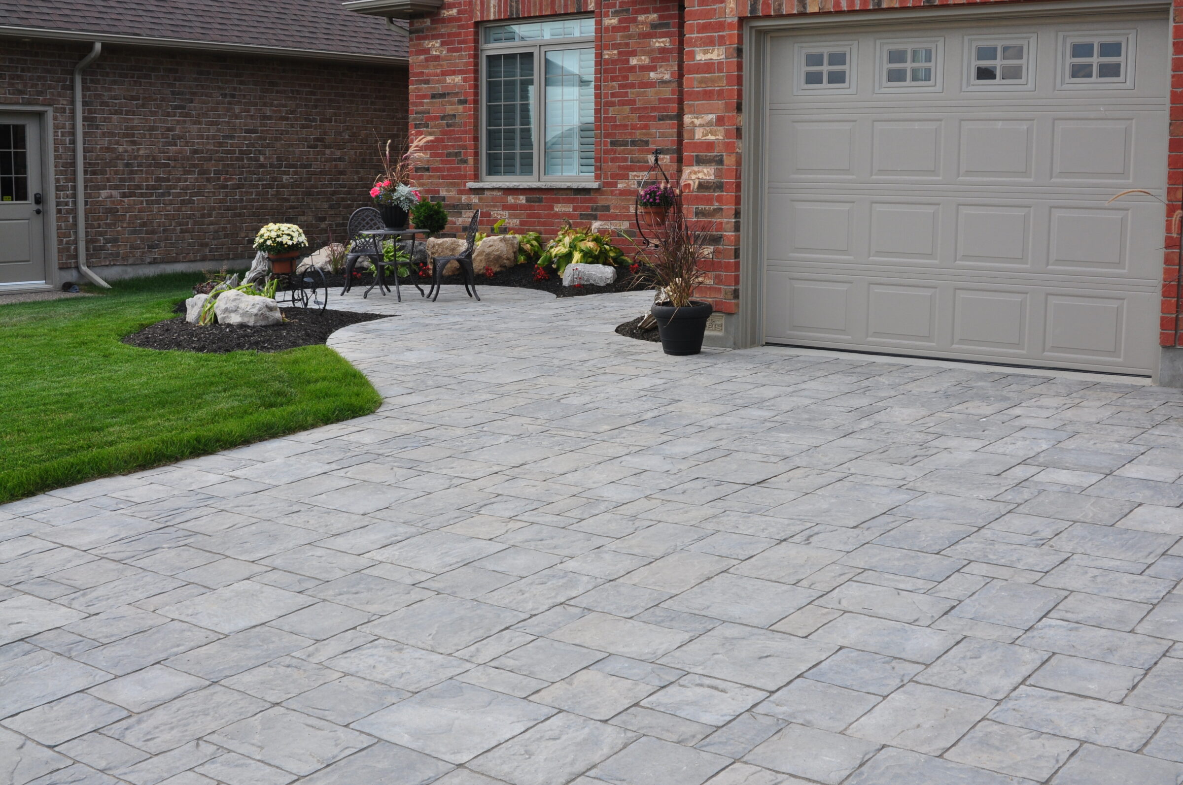 Brick house with a gray garage door, paved driveway, and landscaped garden featuring chairs, plants, and rocks. Cozy and inviting outdoor setting.