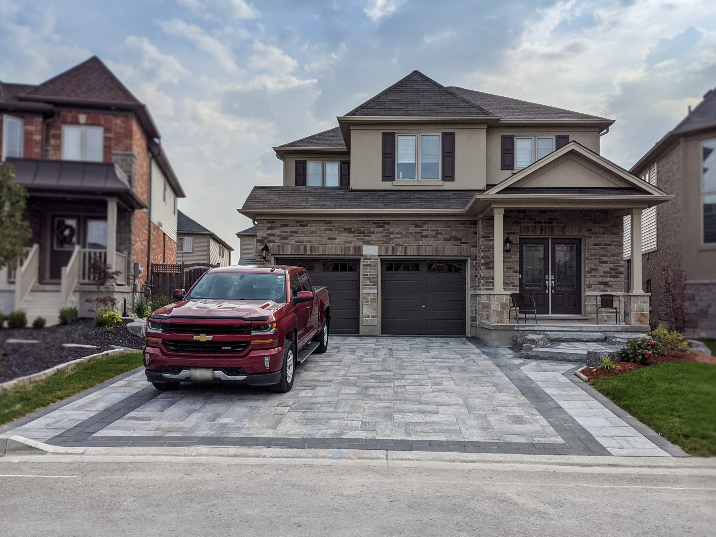 A modern suburban house with a red truck parked on a patterned driveway. The neighborhood features similar style homes under a cloudy sky.