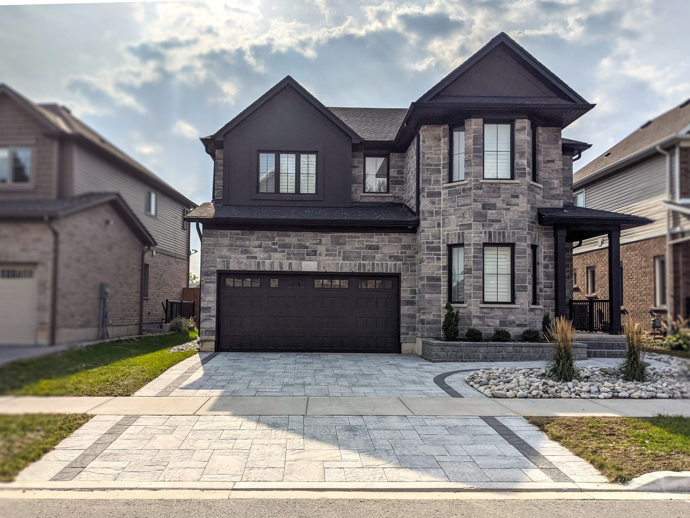 A modern, two-story stone house with a paved driveway and minimal landscaping sits between two similar residential homes under a partly cloudy sky.