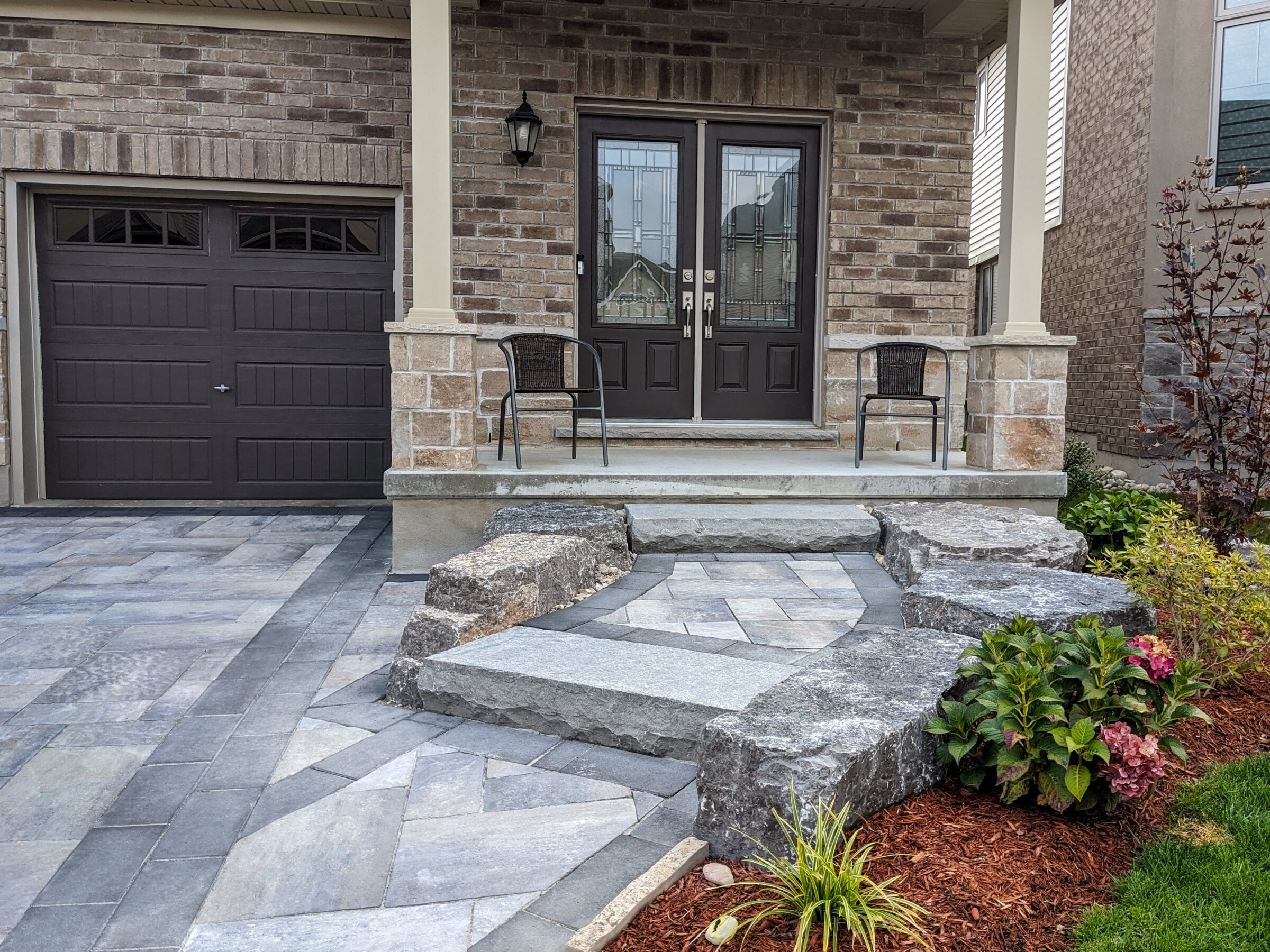 Modern house entrance with dark double doors, geometric stone pathway, manicured bushes, two chairs, a porch, and a garage door on the left.