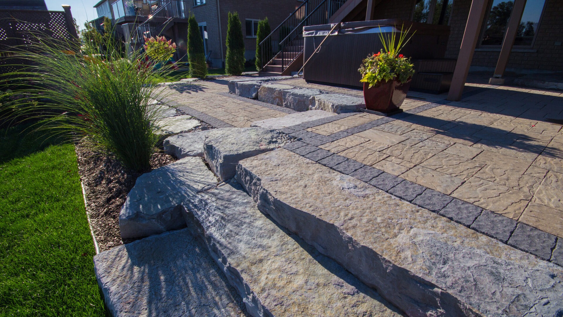 Stone steps and pavers lead to a patio with a hot tub, surrounded by lush greenery and potted plants, in a backyard setting.