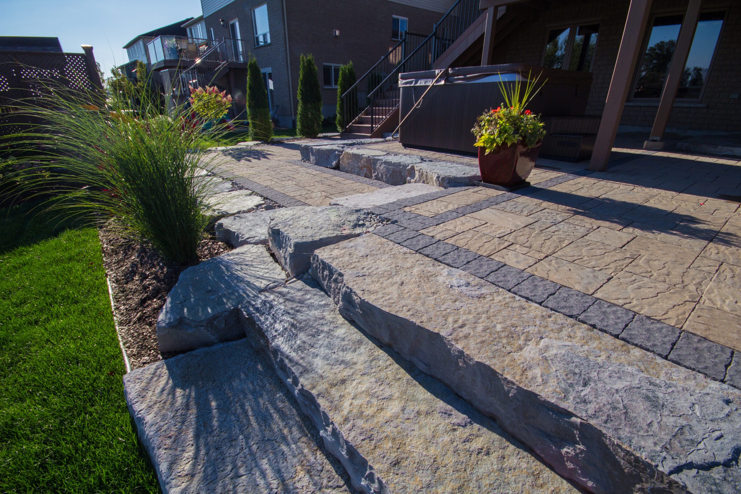 Stone steps lead to a landscaped backyard featuring a hot tub, potted plants, and lush grass beside a modern house.