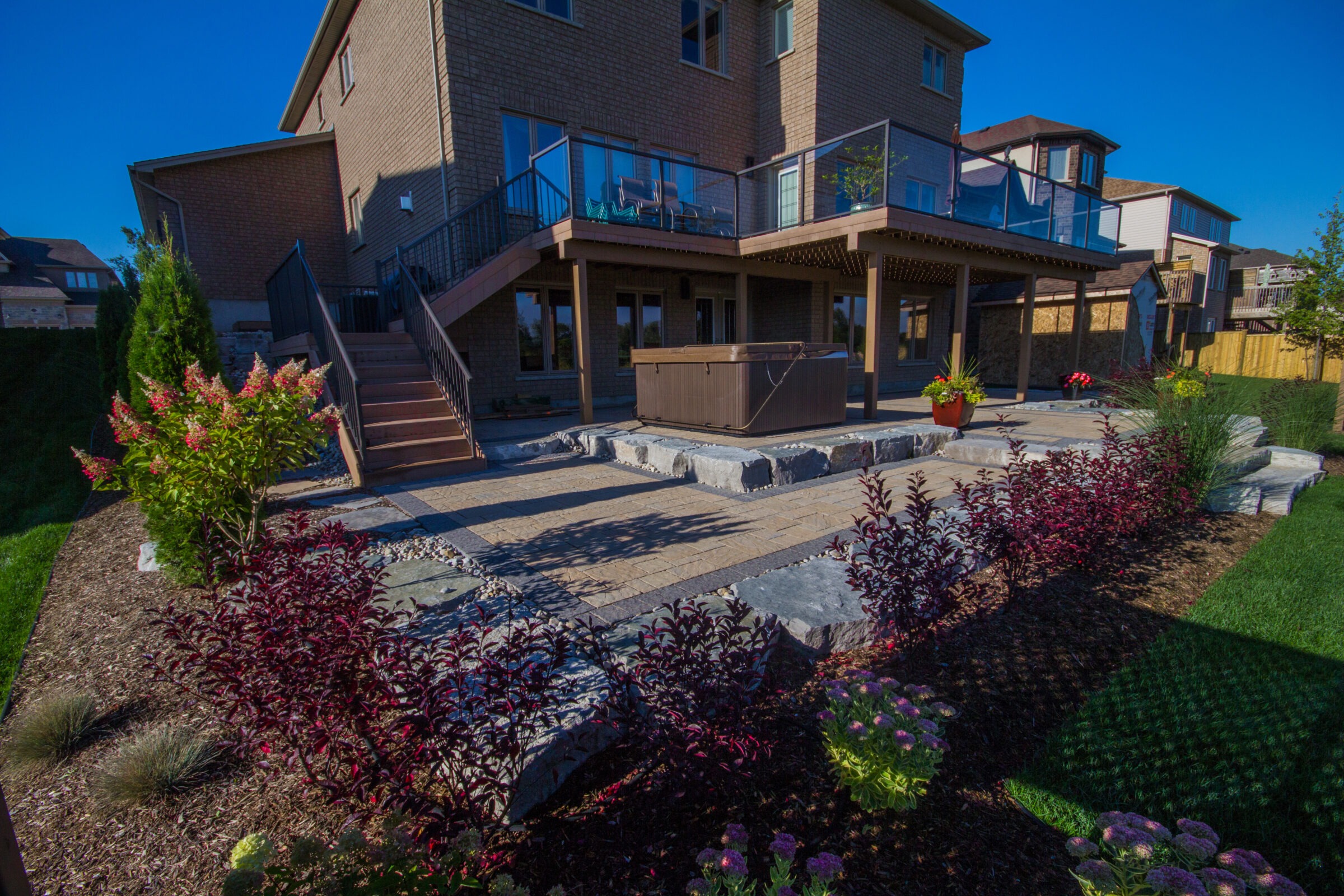 Modern backyard features a multi-level deck with glass railings, hot tub, and landscaped garden showcasing vibrant shrubs and flowers under a clear sky.