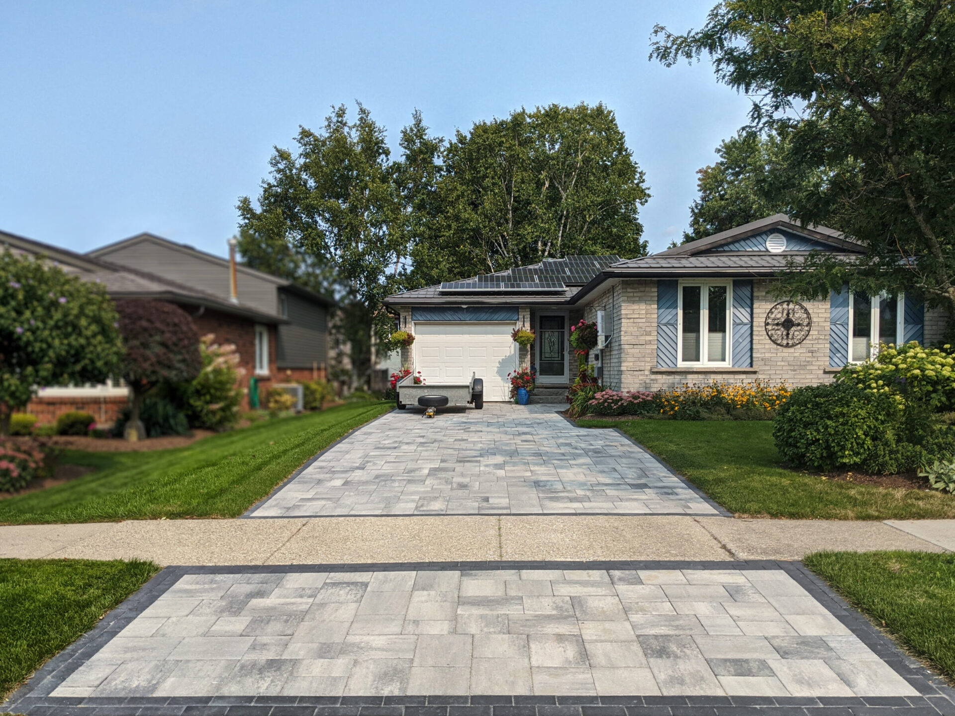 A single-story brick house with well-maintained garden and paved driveway, surrounded by trees, under a clear blue sky. Solar panels are visible.