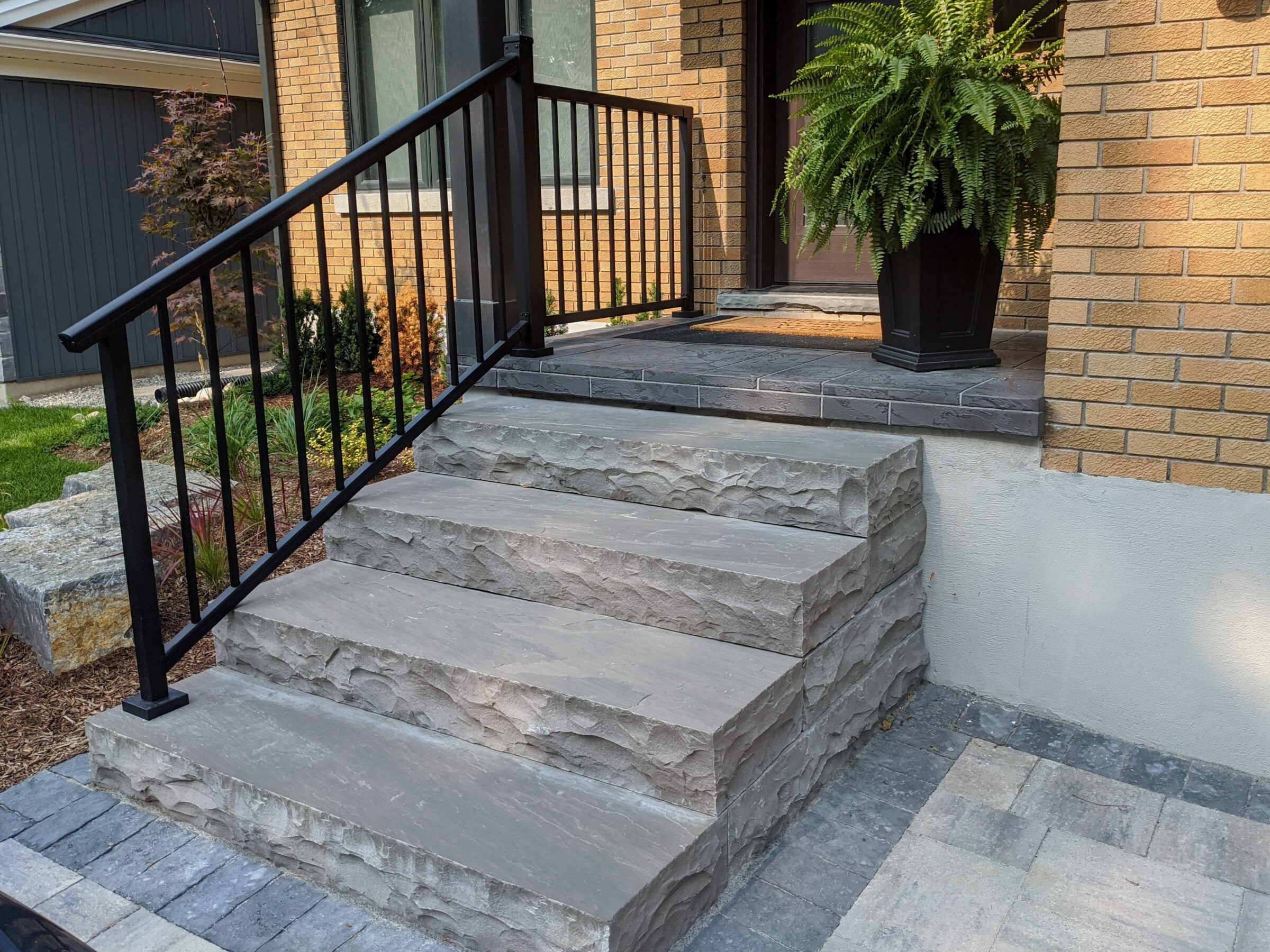 Stone steps leading to a door with a metal railing and a large fern in a pot, surrounded by brickwork.