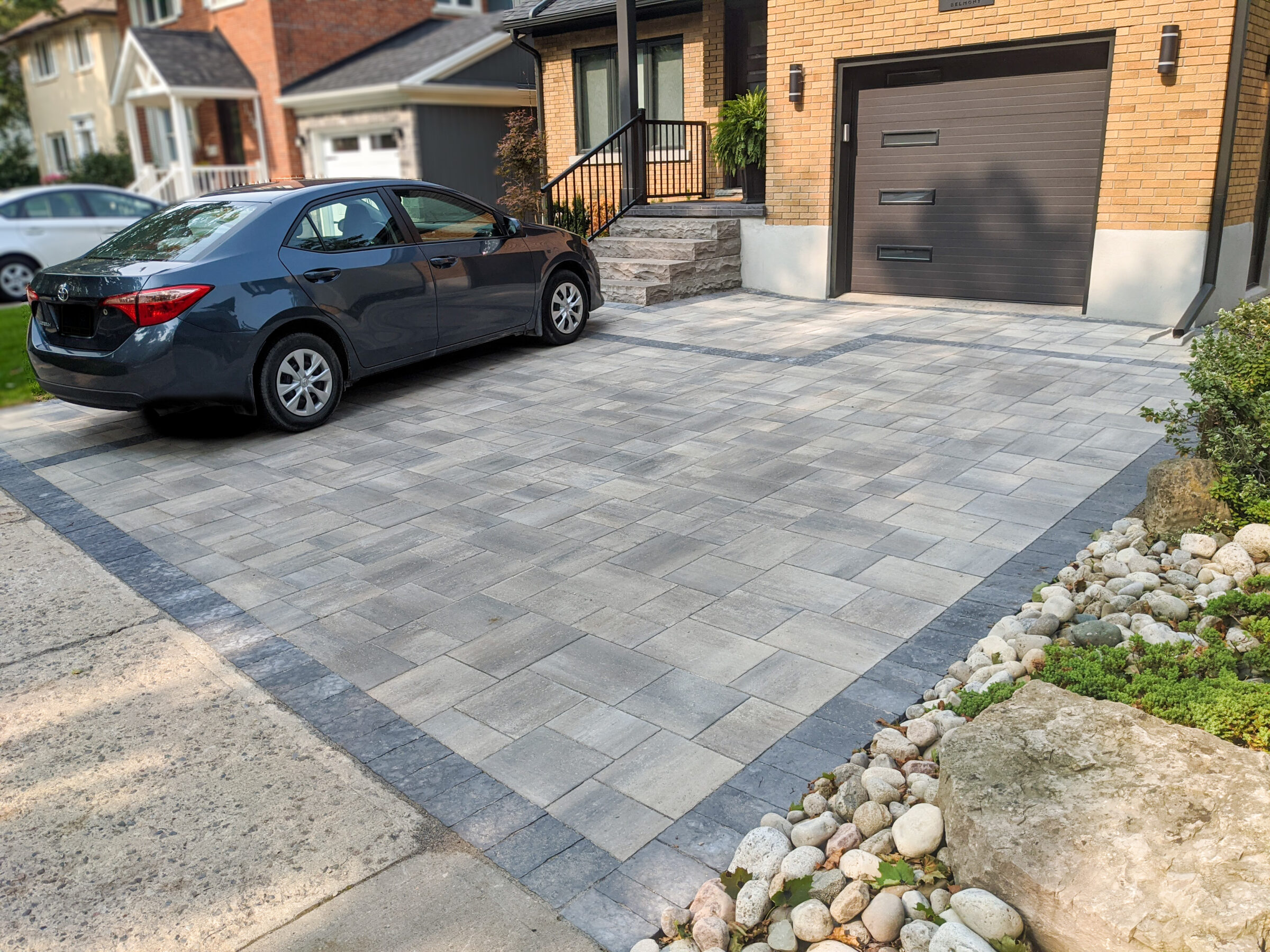 A dark car is parked on a neatly paved driveway beside a brick house, with a small rock garden on the side.