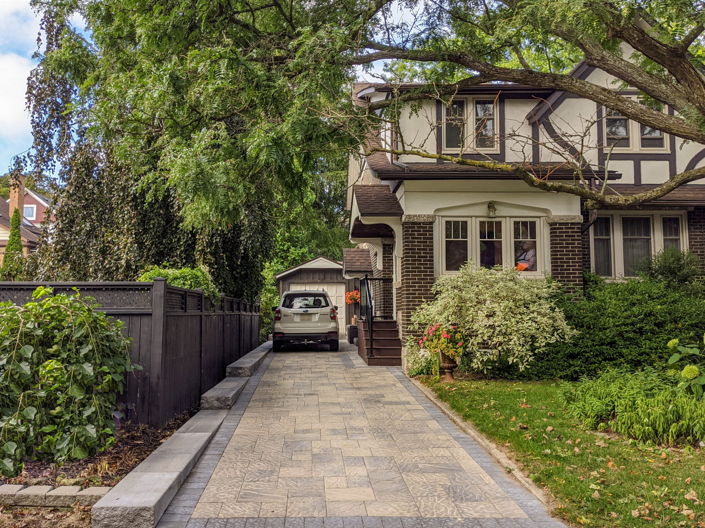 A charming tudor-style house with a stone driveway, lush greenery, and a car parked near a detached garage under a large tree.