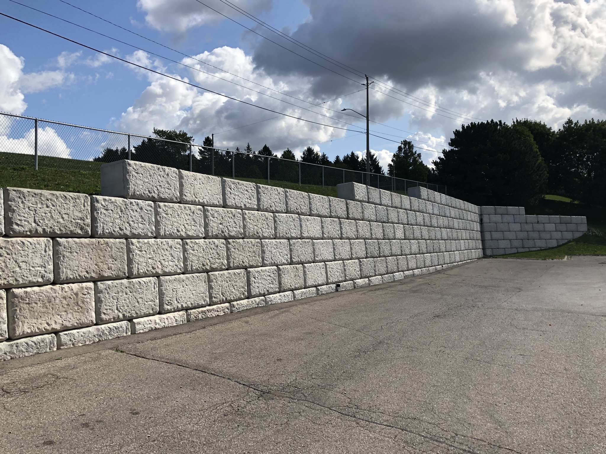 Large concrete retaining wall beside an empty paved area, with a fence and trees under a cloudy sky. No people or landmarks visible.