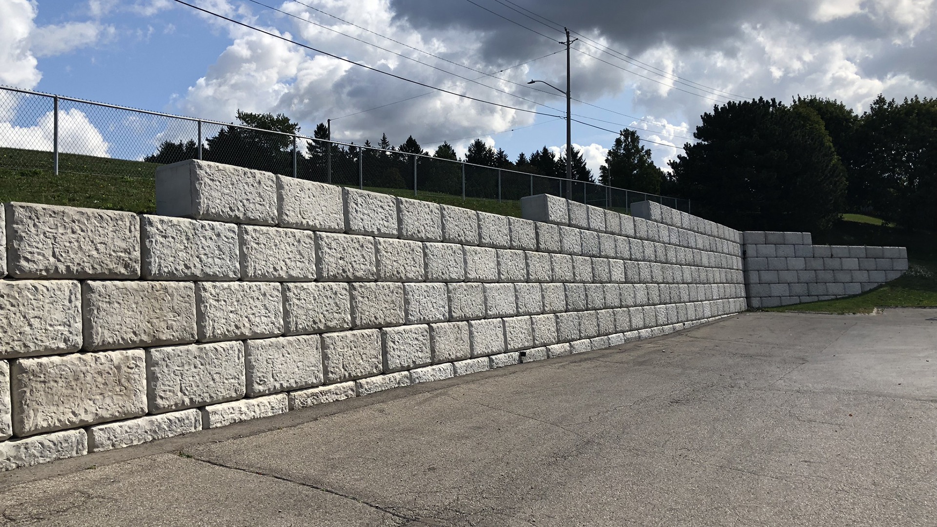 A segmented stone retaining wall with a chain-link fence, surrounded by grass and trees under a partly cloudy sky.