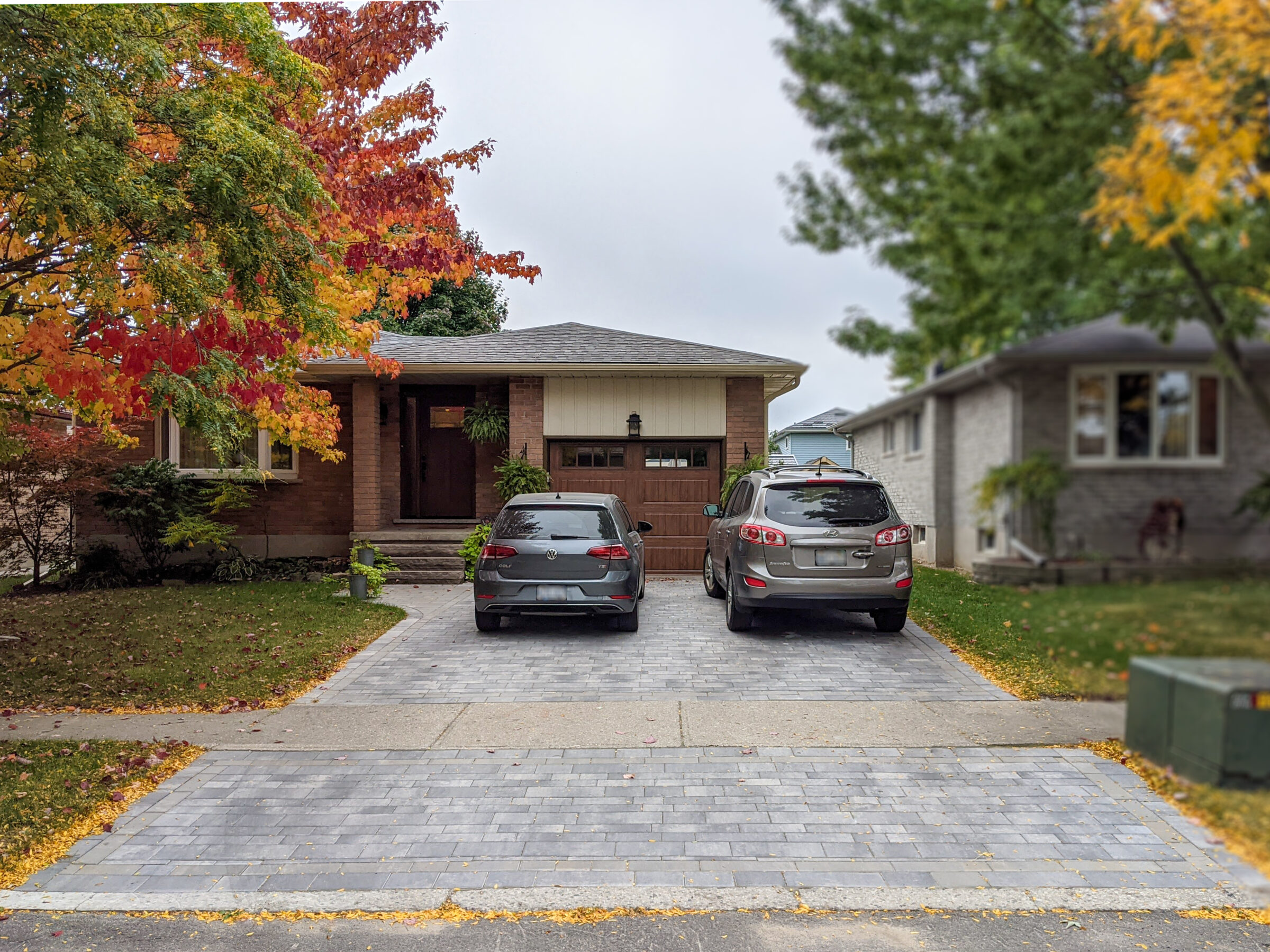 A suburban house with a two-car driveway is surrounded by autumnal trees with red and green foliage, set in a residential neighborhood.
