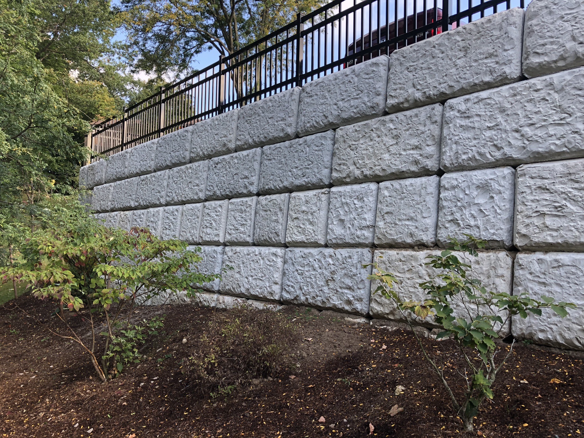 A textured stone retaining wall supports a black metal fence, with leafy trees and shrubs at the base, under a clear sky.