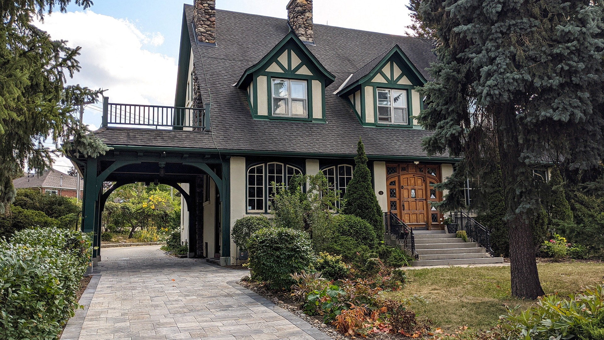 Charming Tudor-style house with green trim, stone chimney, and landscaped garden, featuring a curved driveway and entrance archway, surrounded by trees.