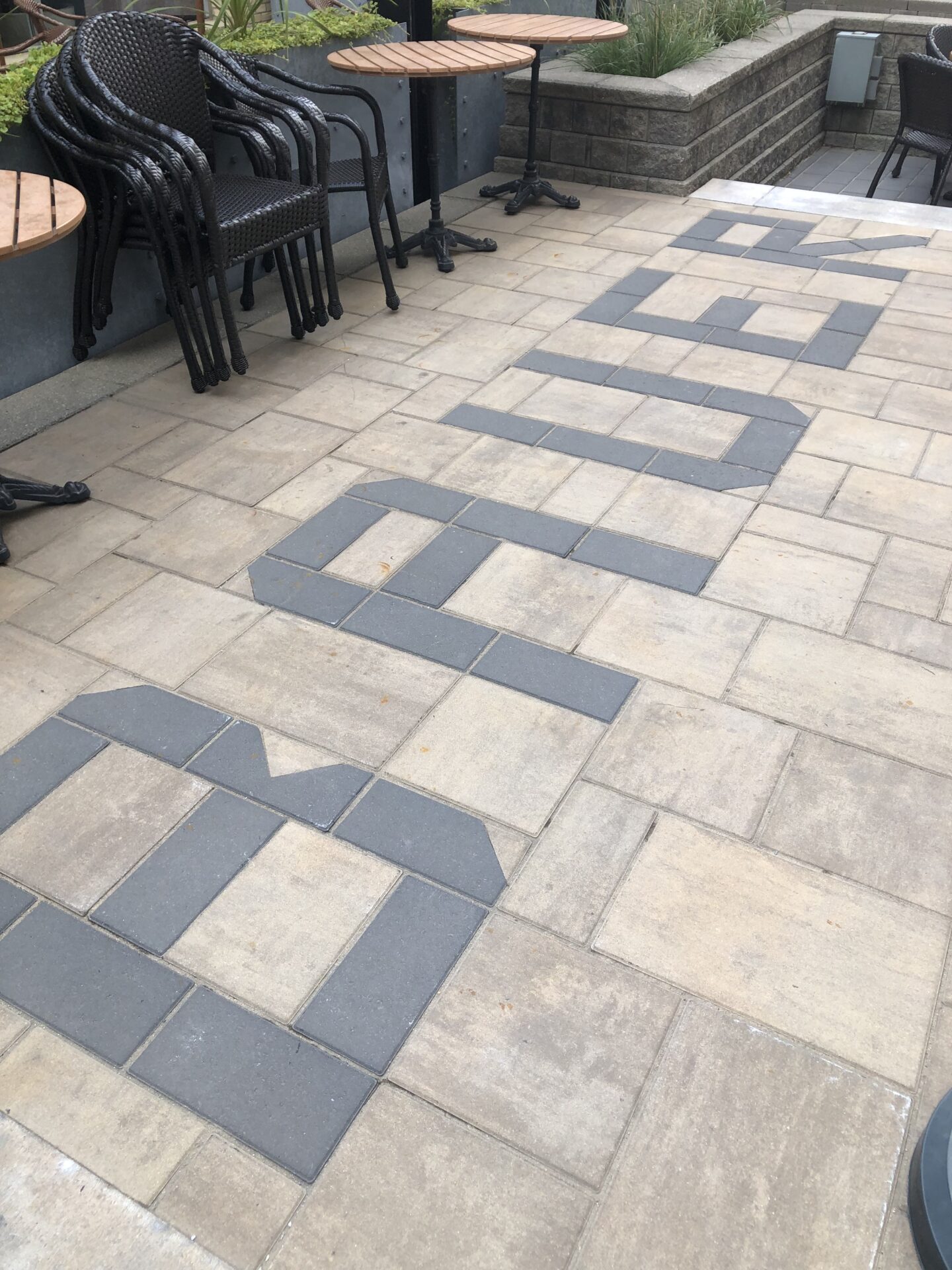 Outdoor patio features wicker chairs, round tables, brick flooring with large letters. Green plants in background, no recognizable landmarks or historical buildings visible.