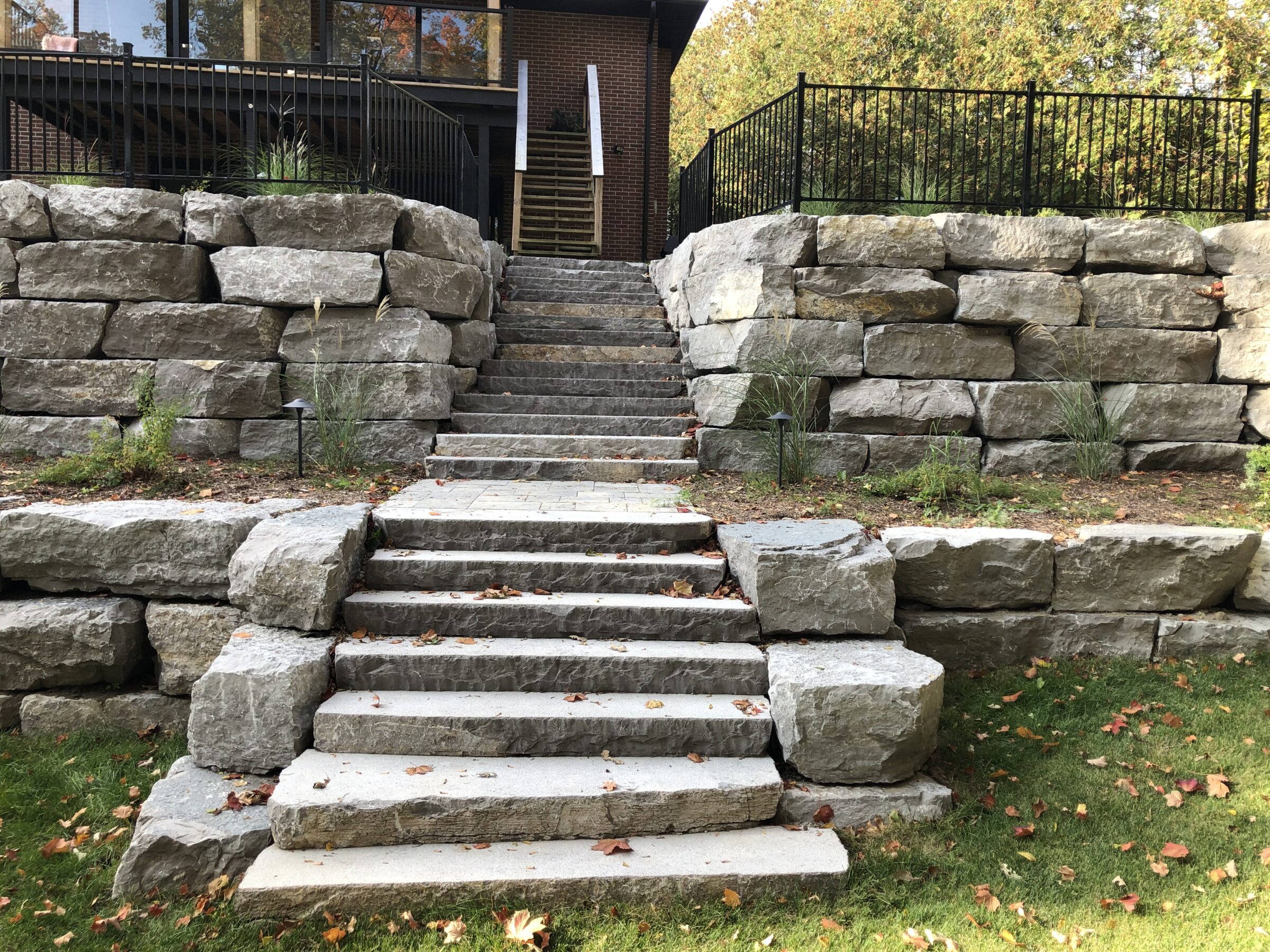 Stone steps lead upward between large rock walls, surrounded by autumn leaves and greenery, ending at a modern brick building with black railings.