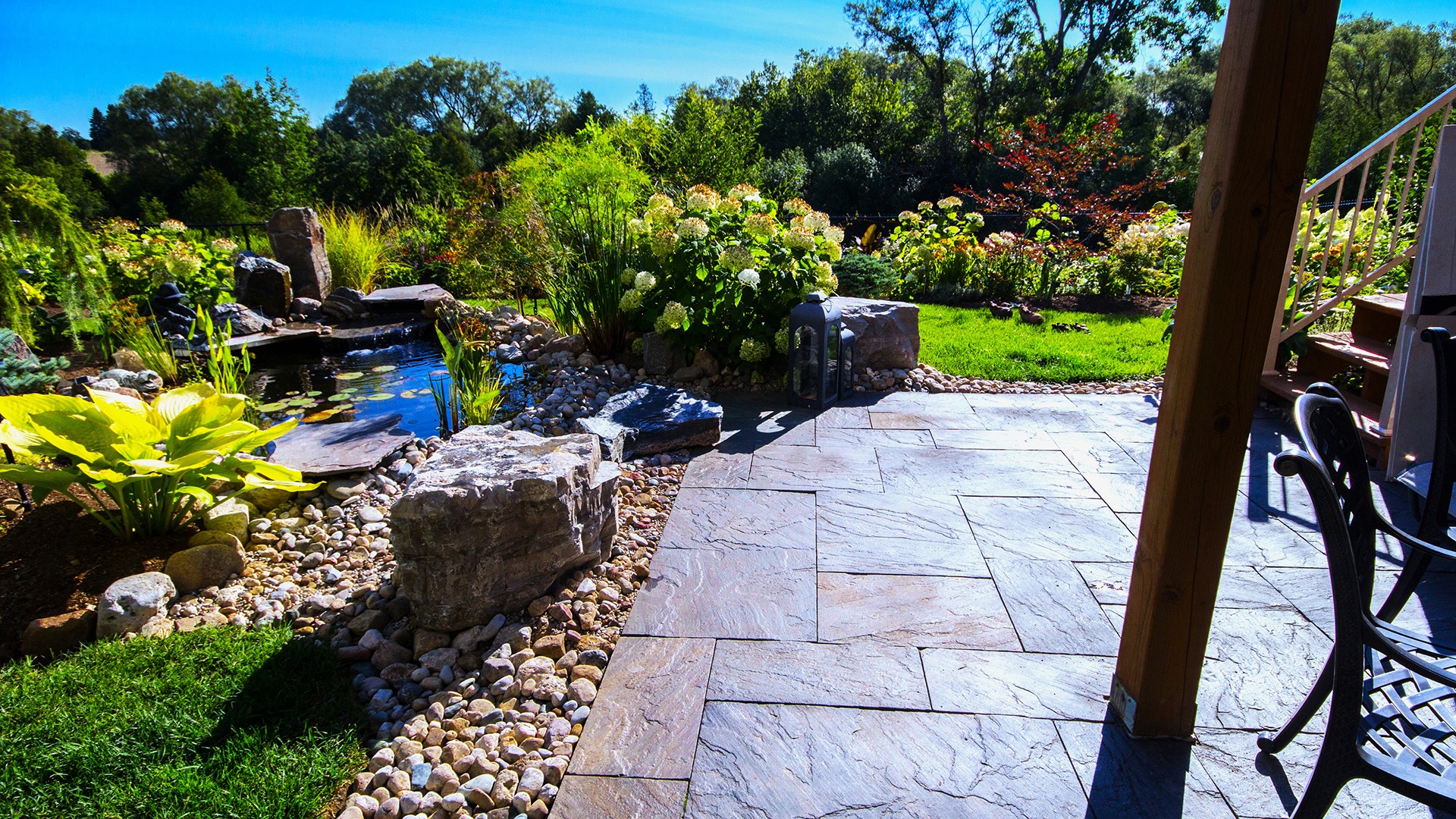 A tranquil garden features a stone patio, lush greenery, a small pond, and a lantern, under a clear blue sky.