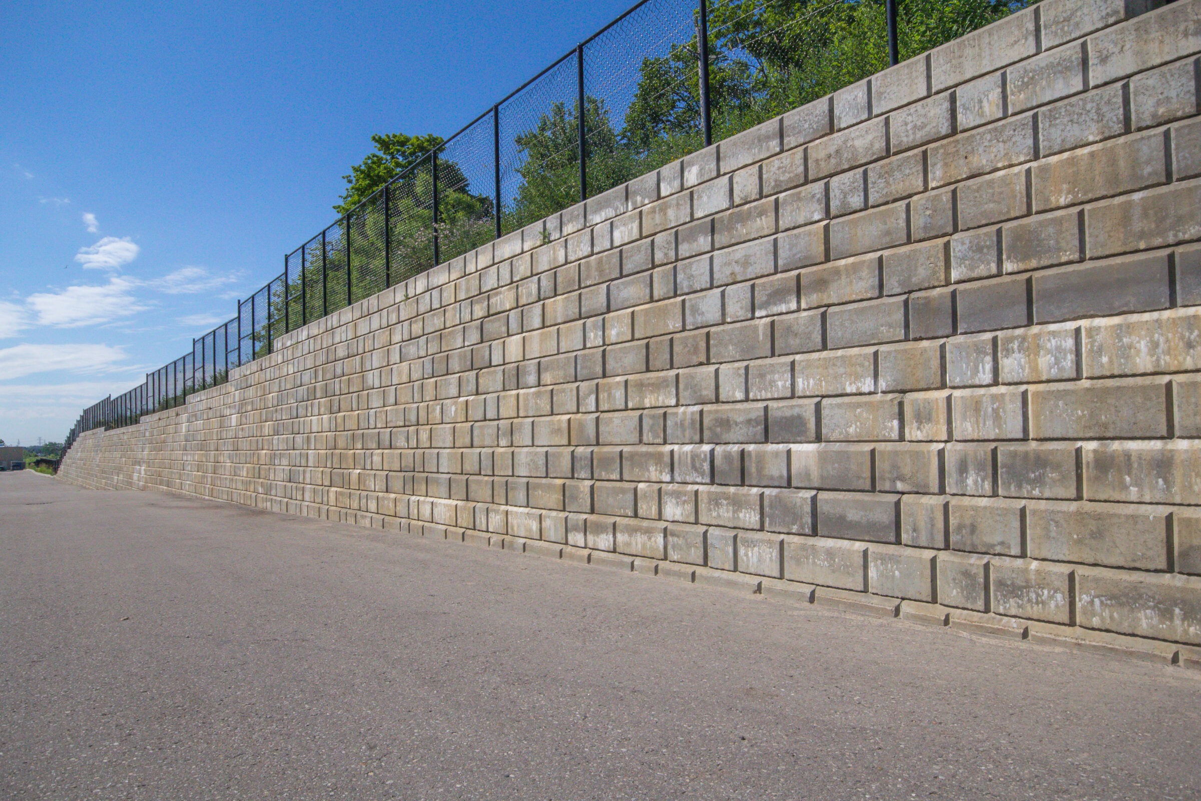 A tall, solid brick retaining wall with a metal fence on a clear day, bordered by trees and an empty paved area.