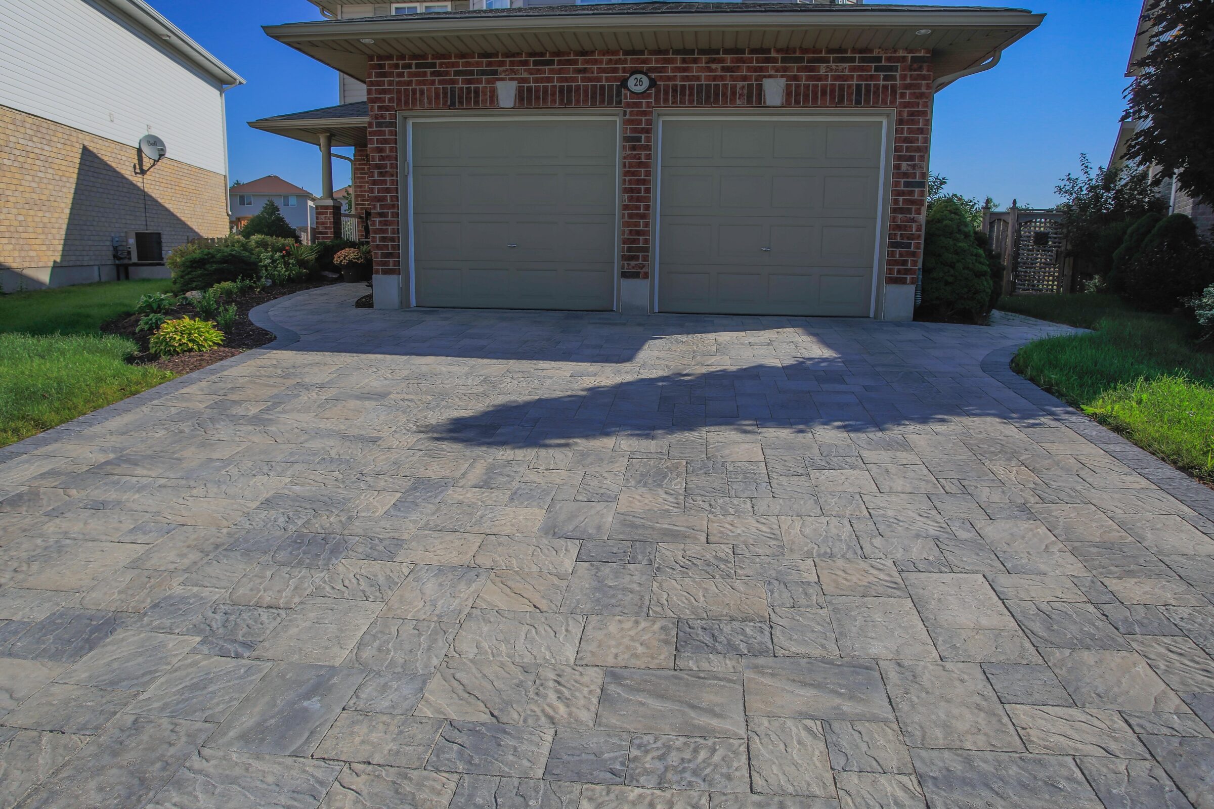 Brick house with a dual garage and paved driveway, surrounded by green lawn and shrubs on a sunny day with a clear sky.