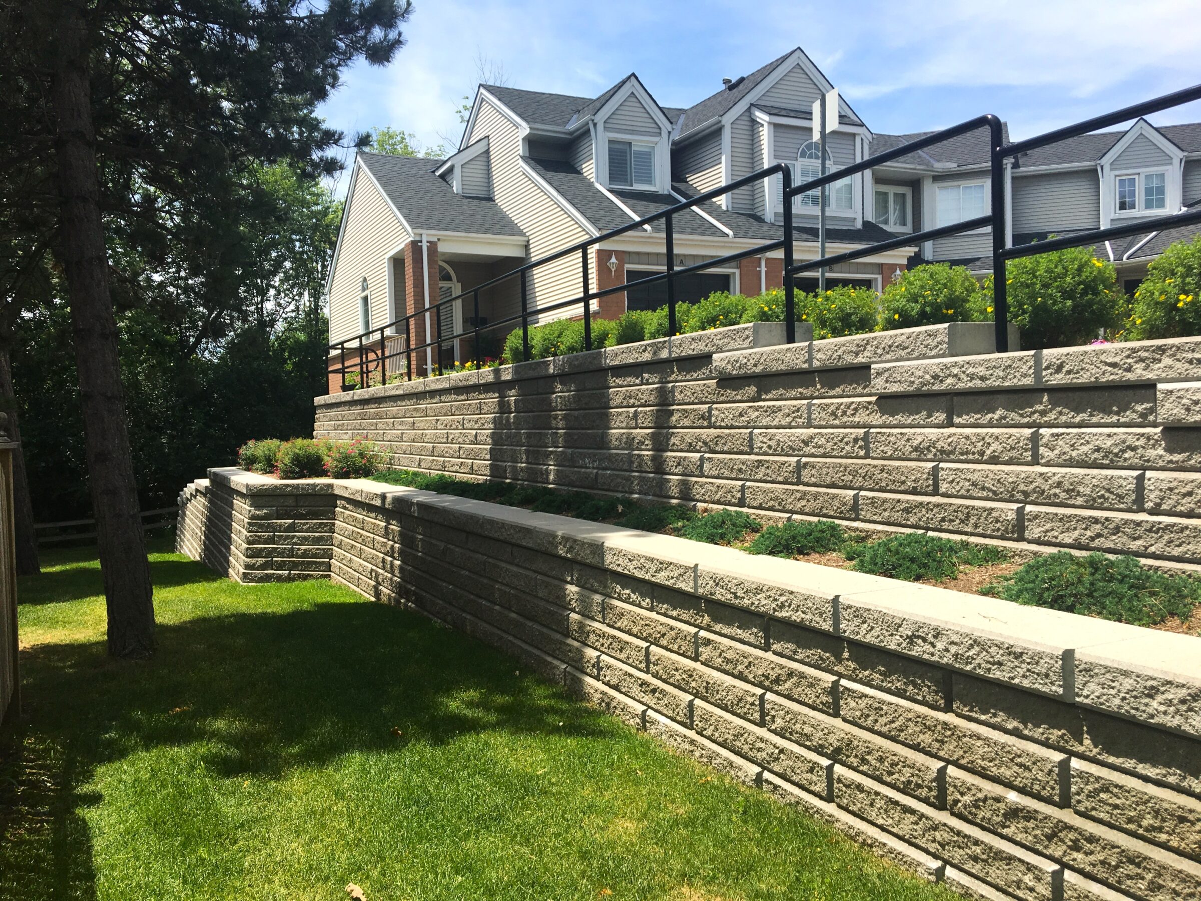 Two-story houses with gray roofs sit behind tiered stone retaining walls, surrounded by green grass and trees under a clear blue sky.