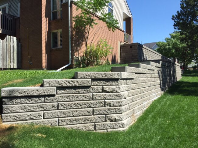 A brick house with a multi-tiered stone retaining wall, lush green grass, and a few trees under clear blue sky.