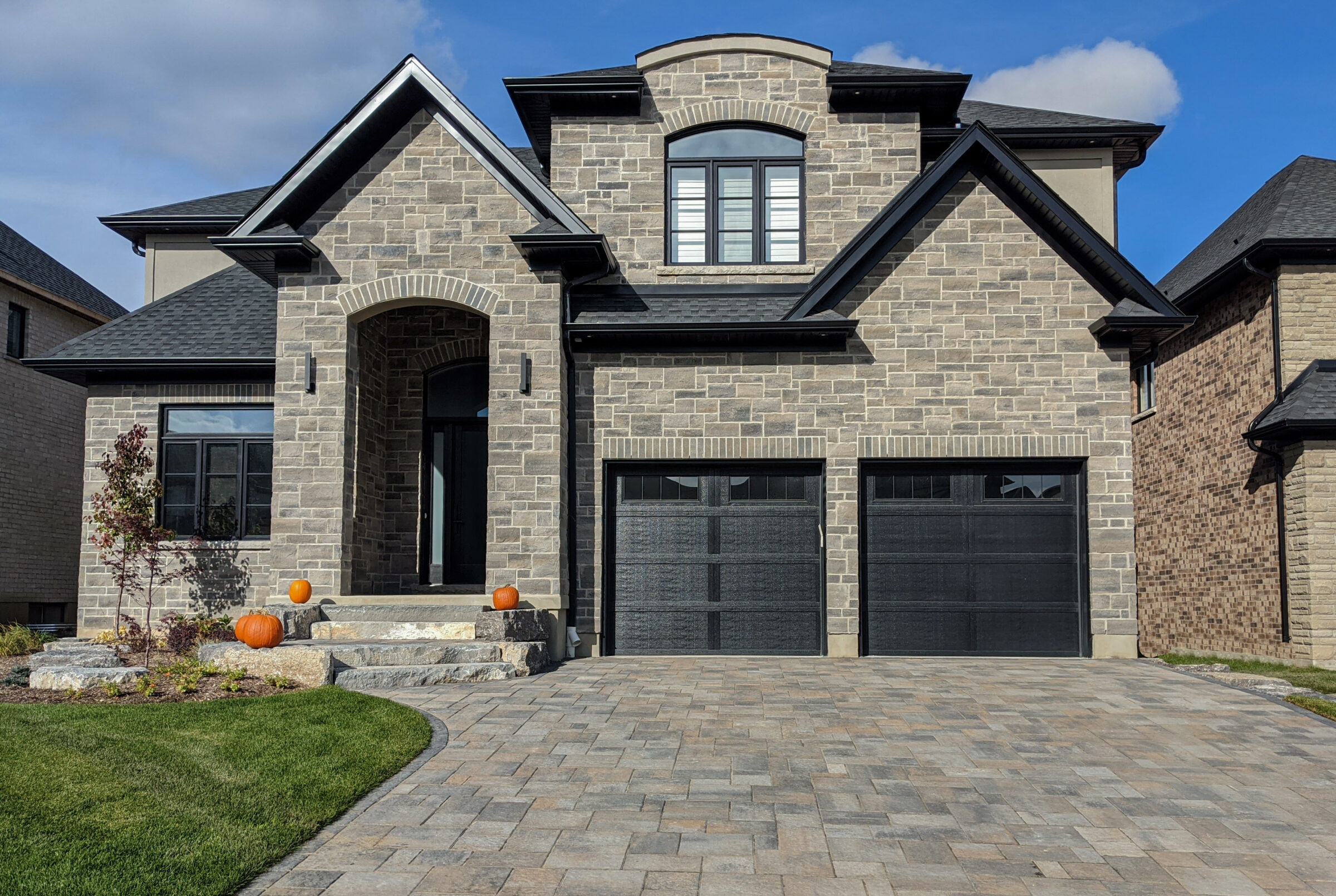 Modern stone house with two black garage doors, arched entrance, and pumpkins on front steps. Well-maintained lawn and clear blue sky.