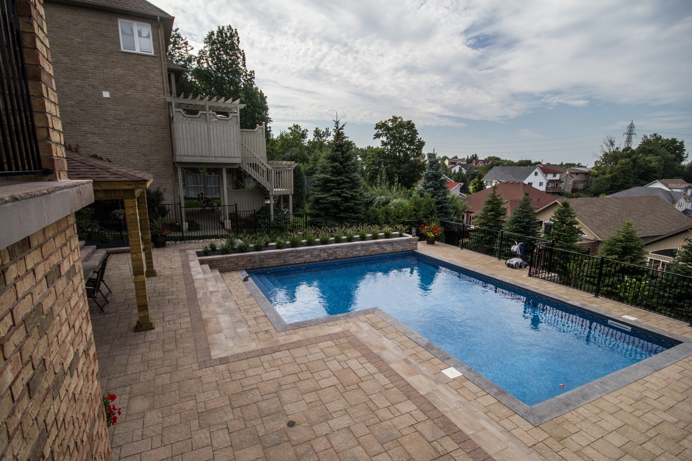 A residential backyard features a large swimming pool, patio with seating, surrounded by lush trees and neighboring houses under a partly cloudy sky.