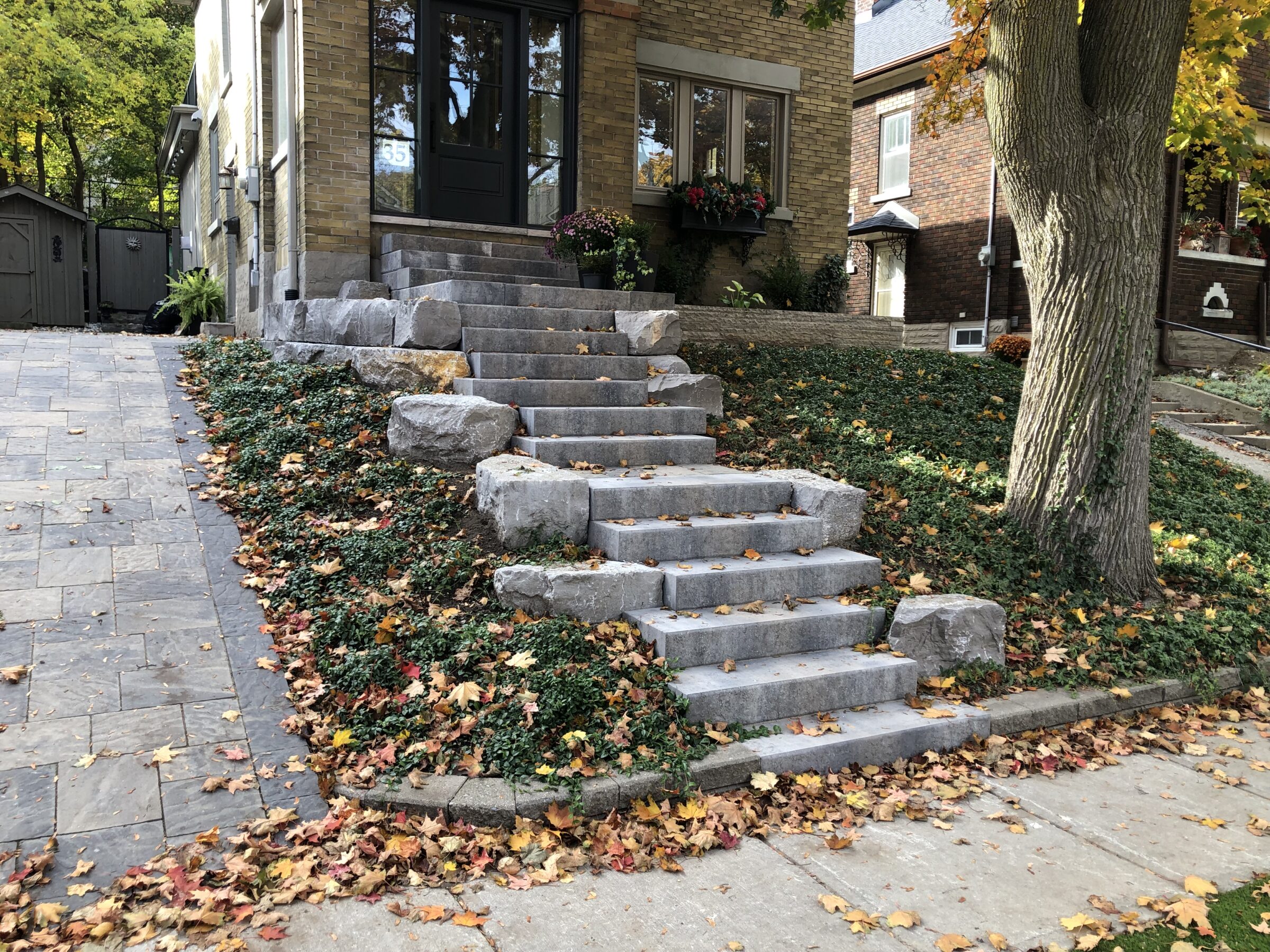 Stone steps lead to a brick house entrance, surrounded by autumn leaves and greenery, with a tree on the right.