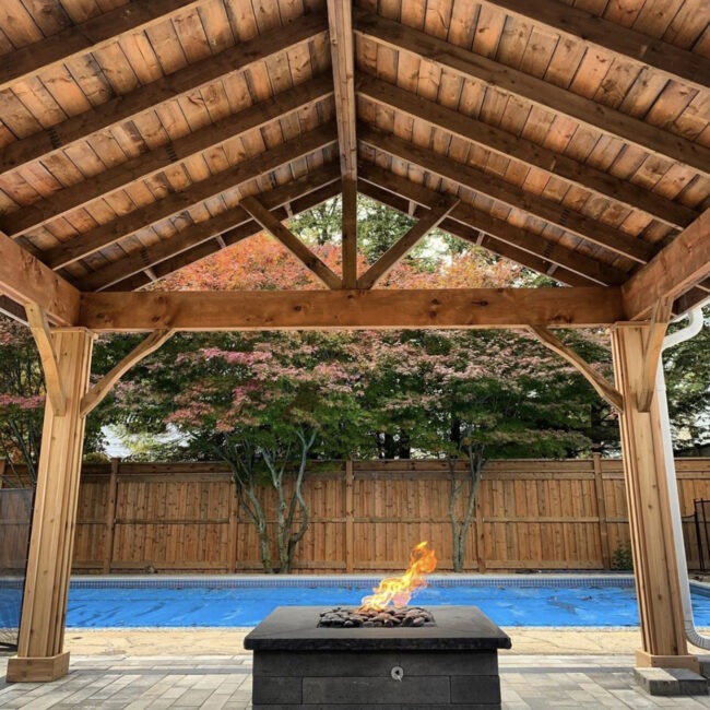 A cozy backyard scene features a wooden pergola, fire pit, covered pool, and vibrant trees, creating a tranquil and inviting atmosphere.