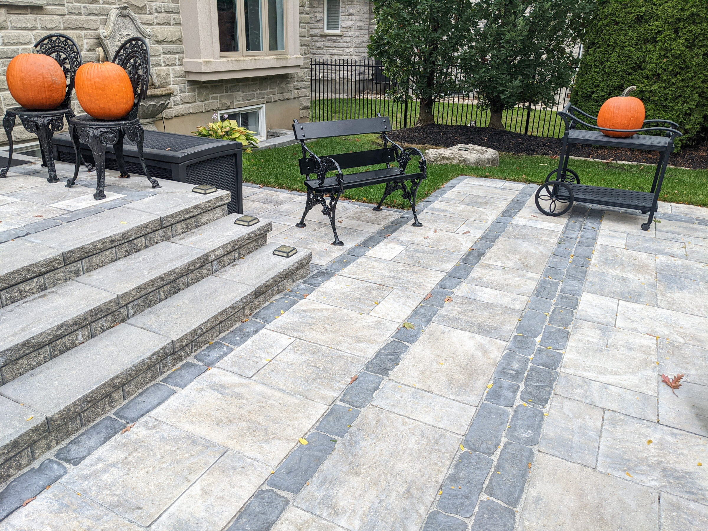 Stone patio with pumpkins on ornate chairs and cart. Bench nearby. Greenery and brick building in the background, no visible landmarks.
