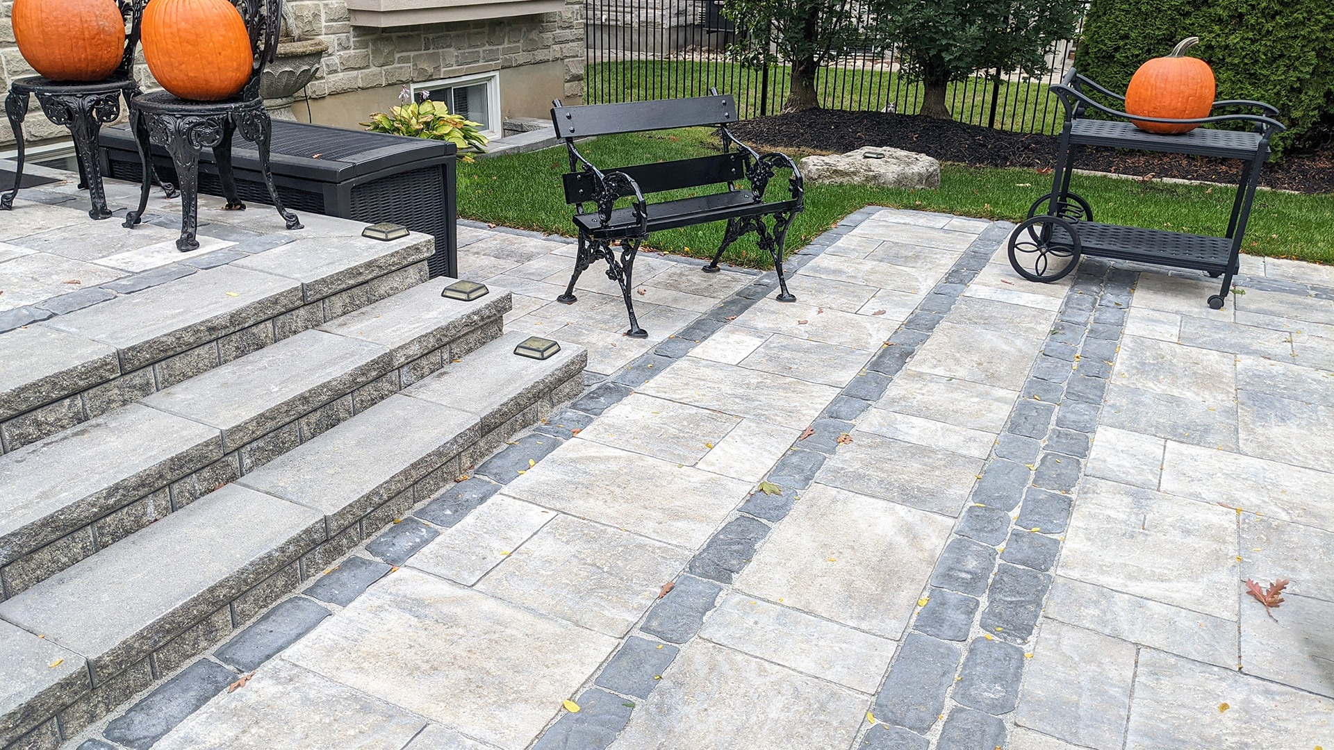 Stone patio with ornate benches, pumpkins, and a cart. Steps lead to a garden area, flanked by greenery and wrought iron fence.