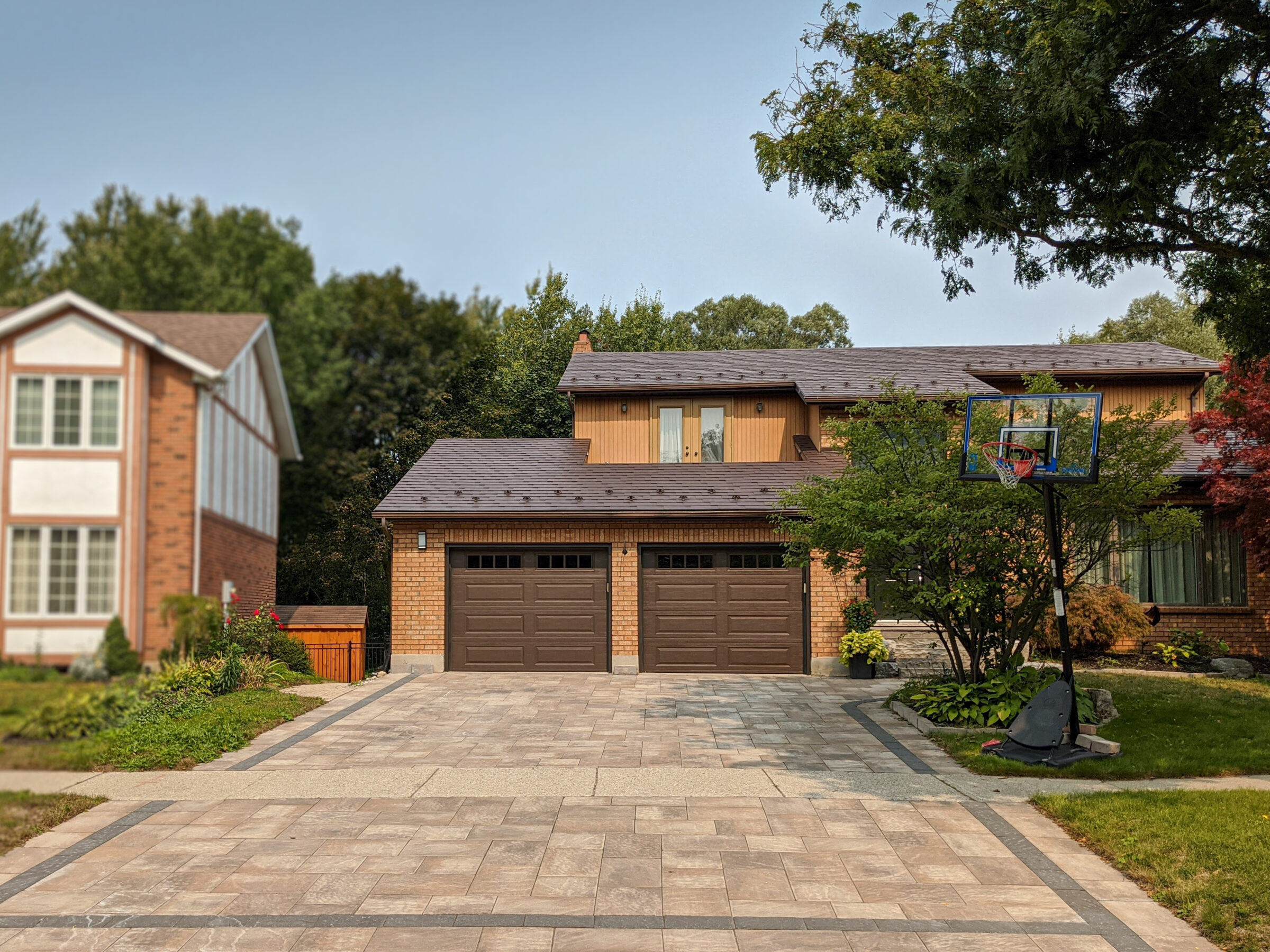 A suburban house with a basketball hoop stands on a spacious driveway, surrounded by trees and greenery, under a clear blue sky.