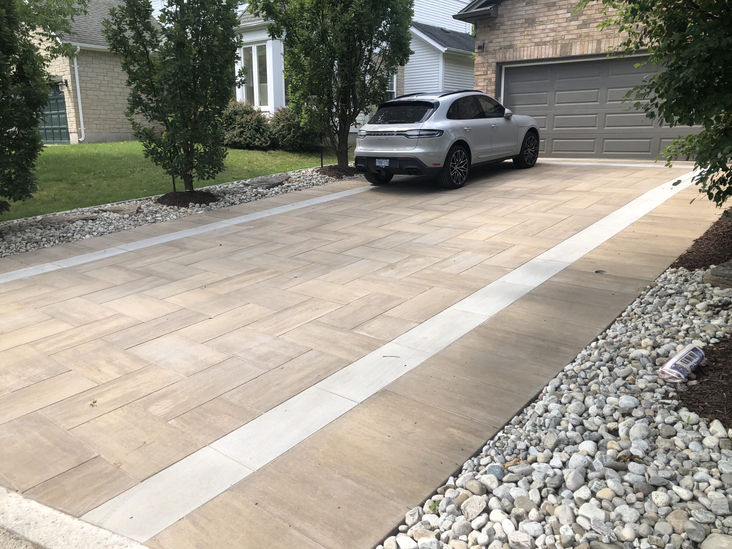 A car is parked on a neatly paved driveway lined with trees and stones outside a brick house with a garage.
