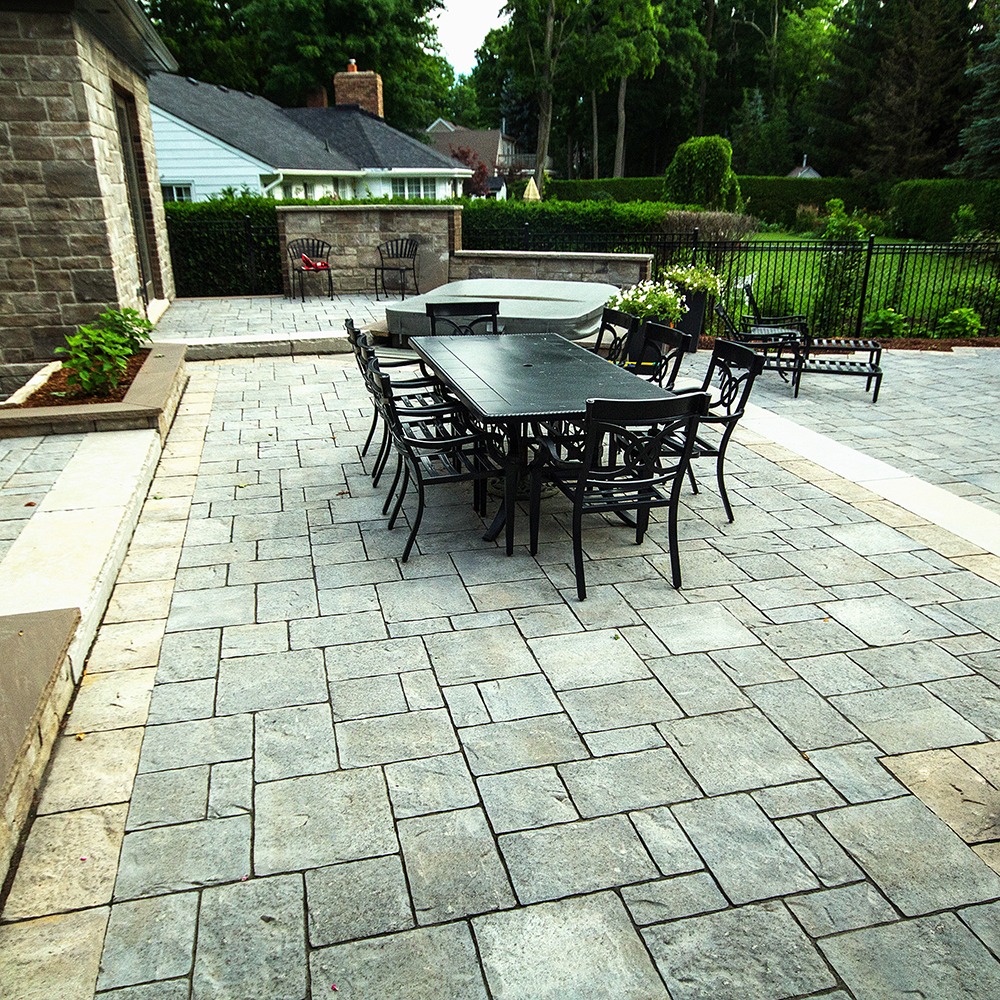 A stone patio with black metal furniture, surrounded by greenery and residential buildings in a quiet suburban setting.