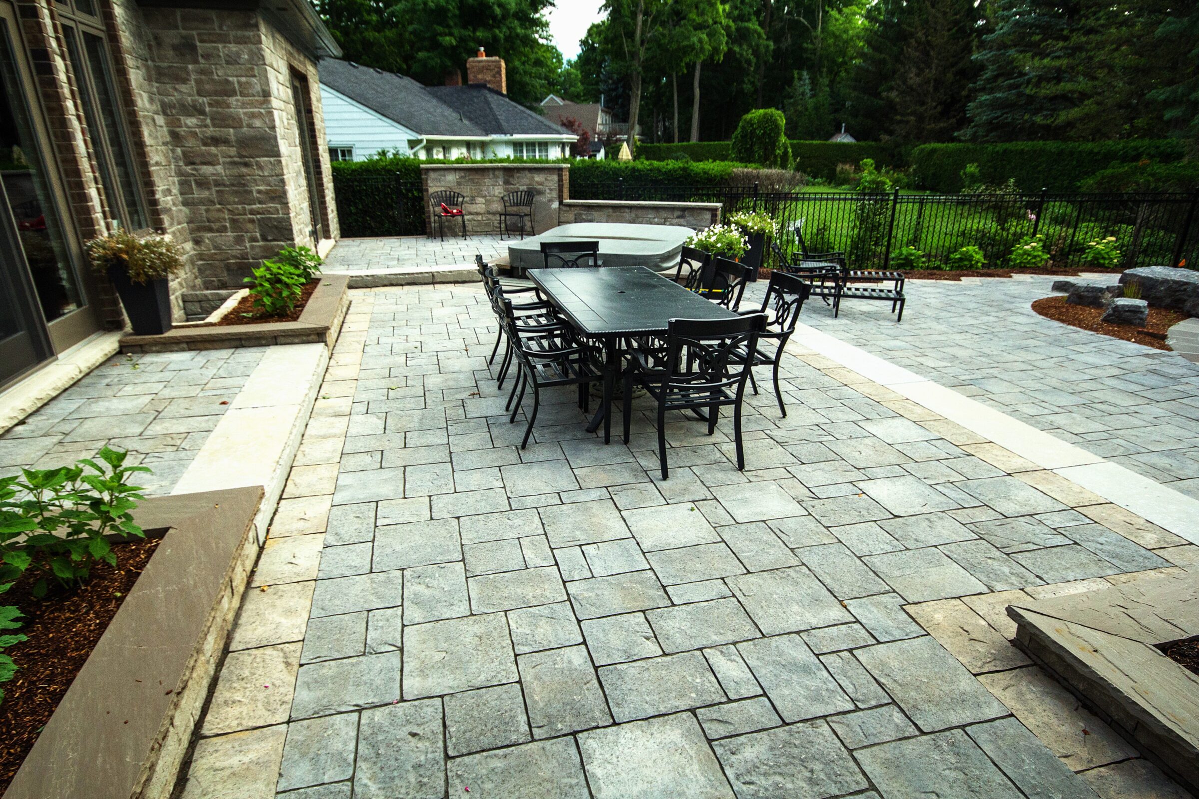 A spacious stone patio with black metal furniture, surrounded by greenery, near a brick building and a residential area.