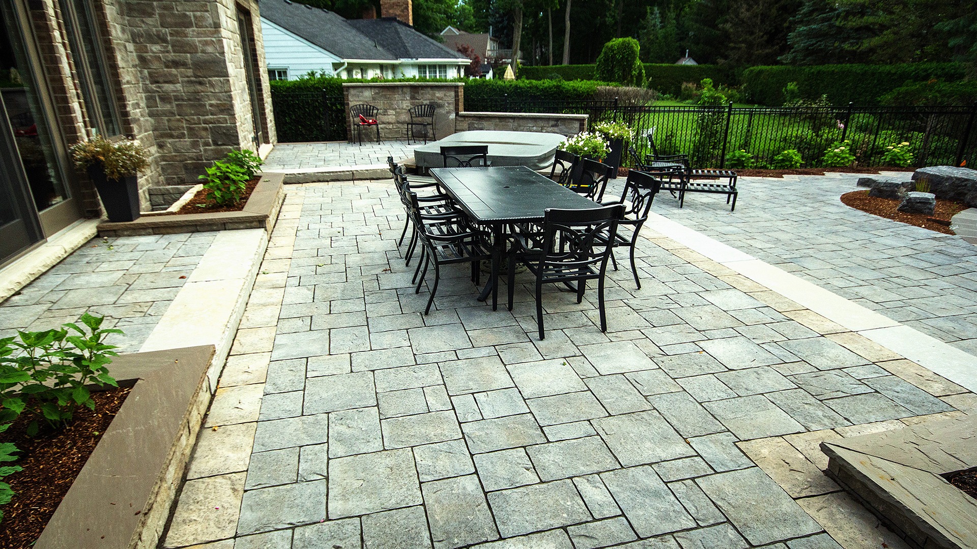 Spacious patio with black metal table and chairs, surrounded by brick walls, lush greenery, and small planters, offering a serene outdoor setting.