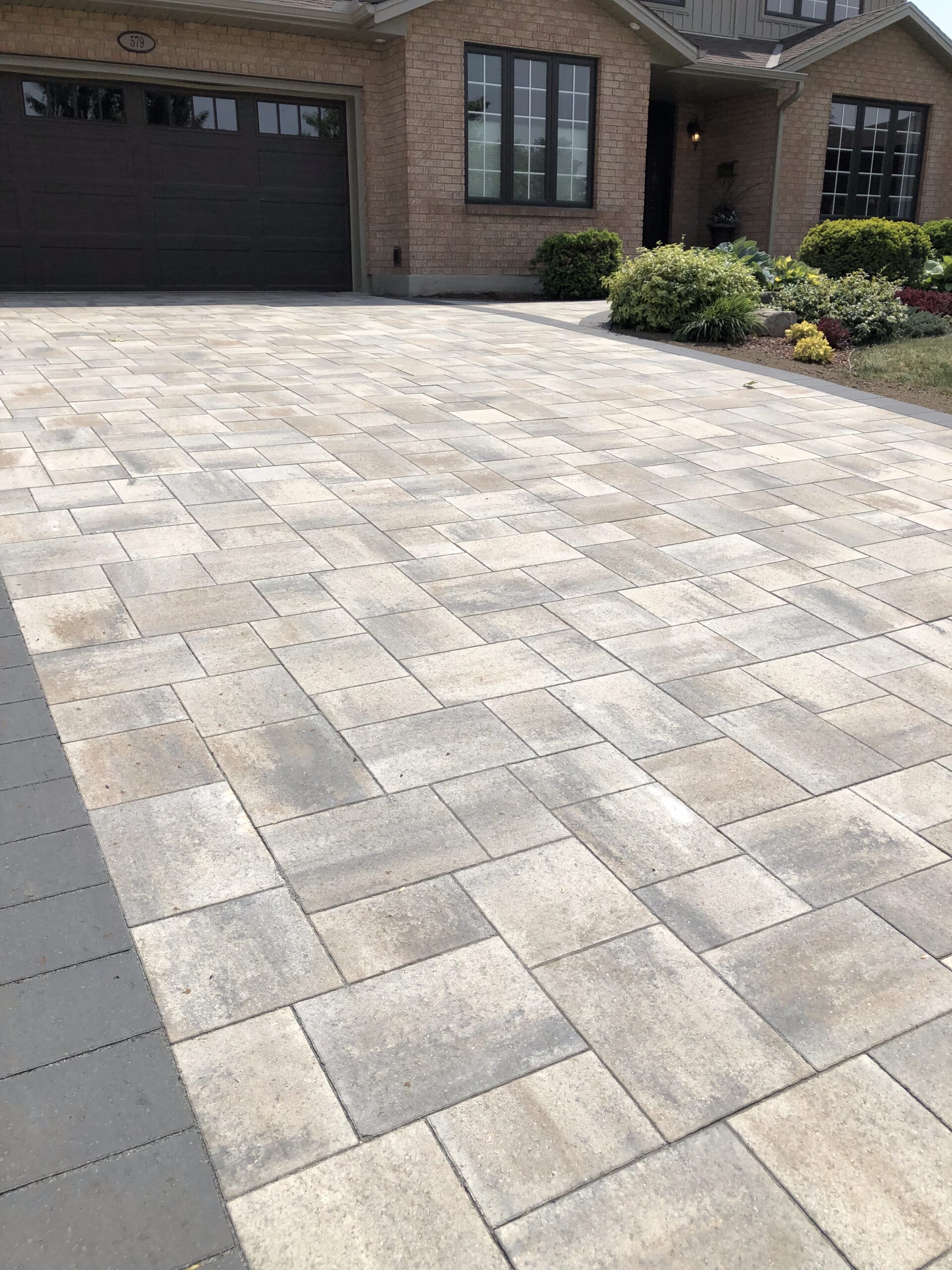 A residential brick house with a dark garage door and neatly landscaped front yard, featuring a newly paved driveway in a geometric pattern.
