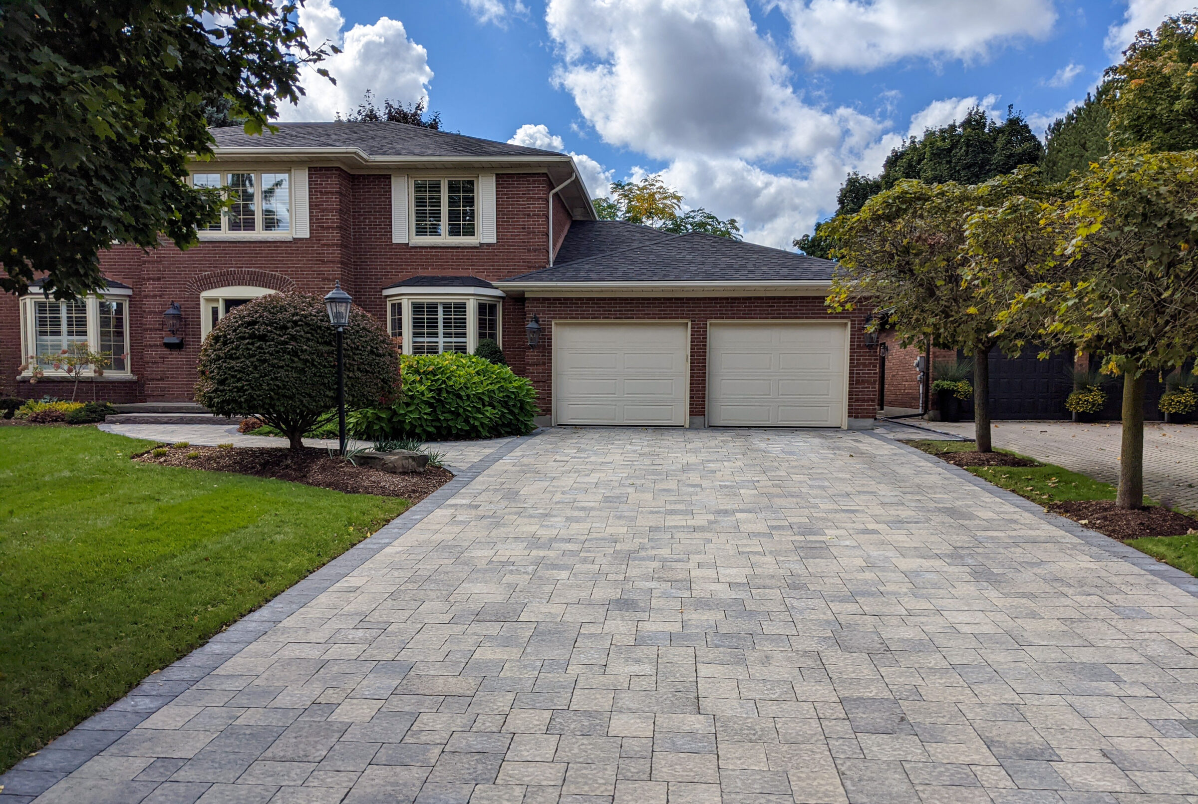 A brick house with a two-car garage, well-maintained landscaping, stone driveway, and trees under a partly cloudy sky.