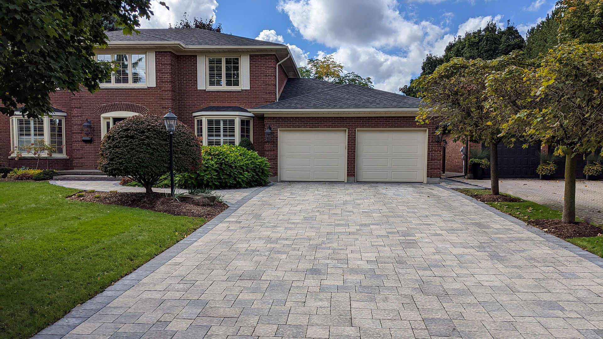 Suburban brick house with a double garage, surrounded by a neat garden, trees, and paver driveway under a partly cloudy sky.