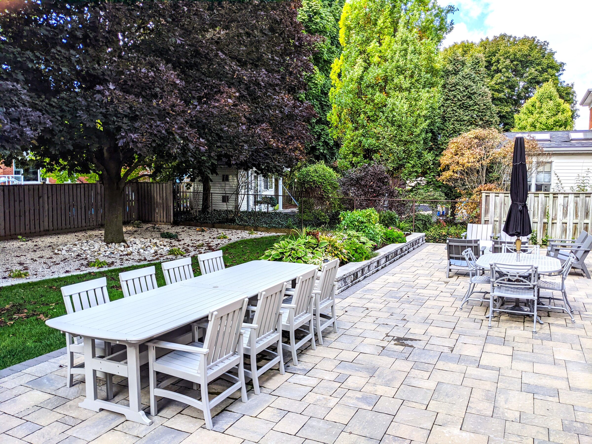 Outdoor patio space with white tables and chairs, surrounded by greenery and trees, adjacent to a house with a fenced yard.