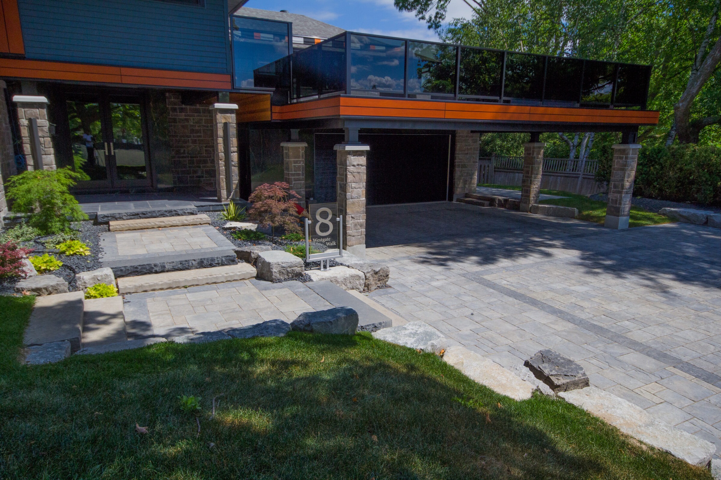 Modern house with stone and glass features, surrounded by landscaped greenery. A sign displays "8 Westgate Walk." Bright, sunny day with clear skies.