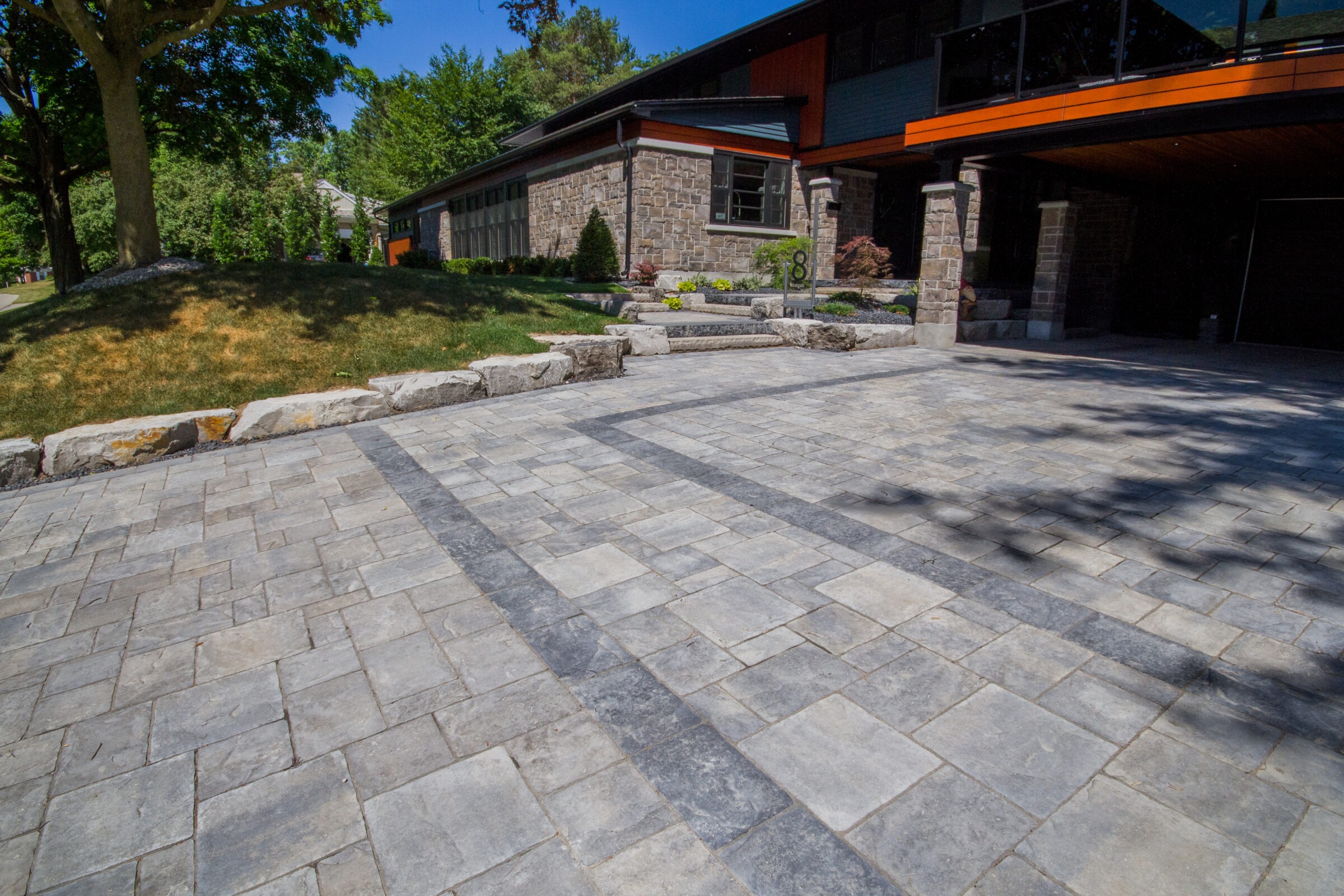 Stone-paved driveway leading to a modern house with stone facade, surrounded by trees and manicured shrubs under a clear blue sky.