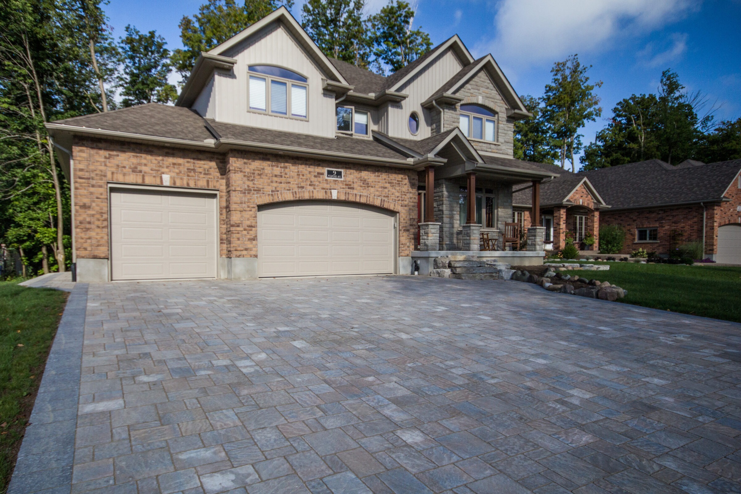A large brick house with two garages, a paved driveway, surrounded by trees under a partly cloudy sky. Quiet residential setting.