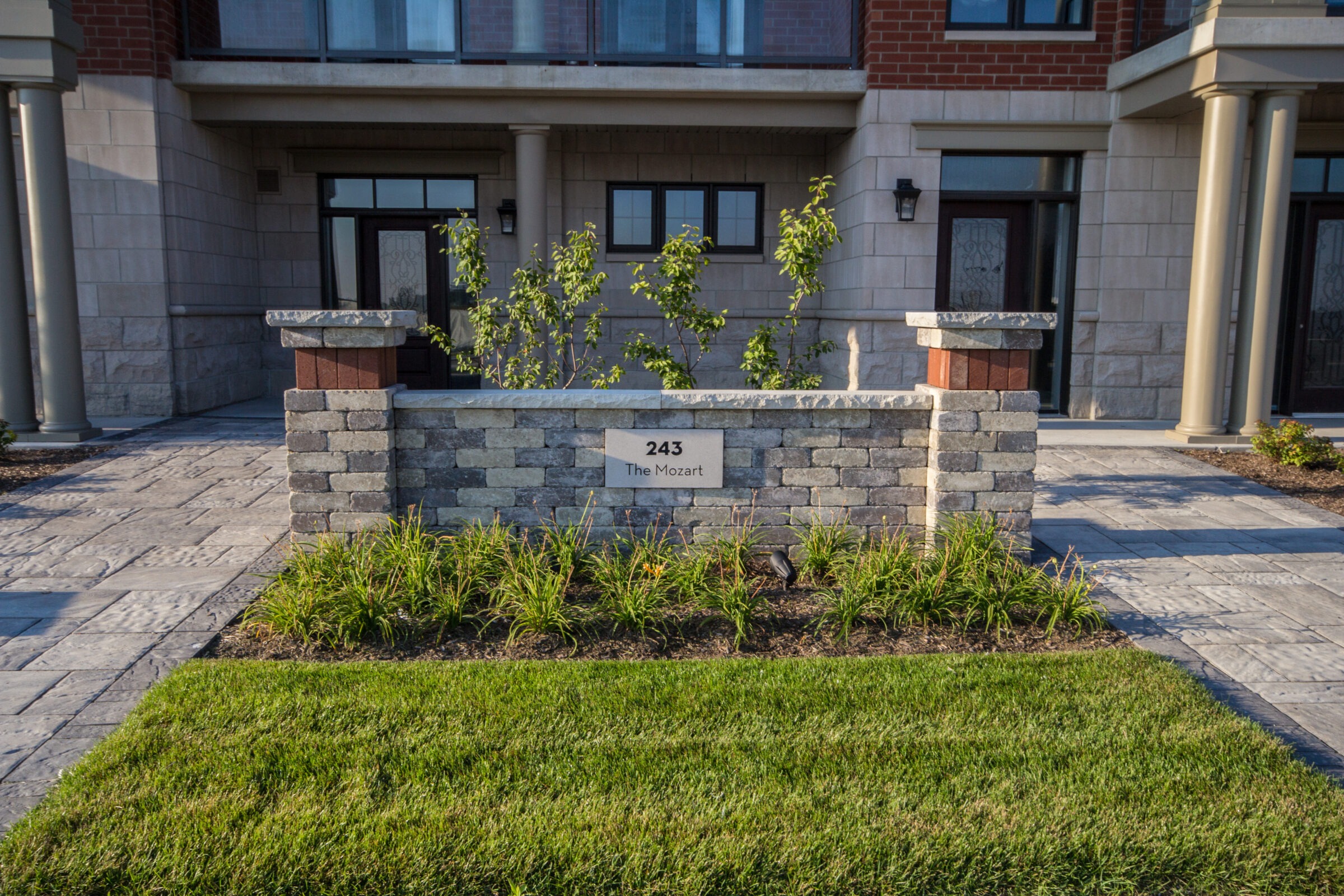 Brick building entrance with pillars and landscaped greenery, featuring a stone sign reading "243 The Mozart" near the doorway.