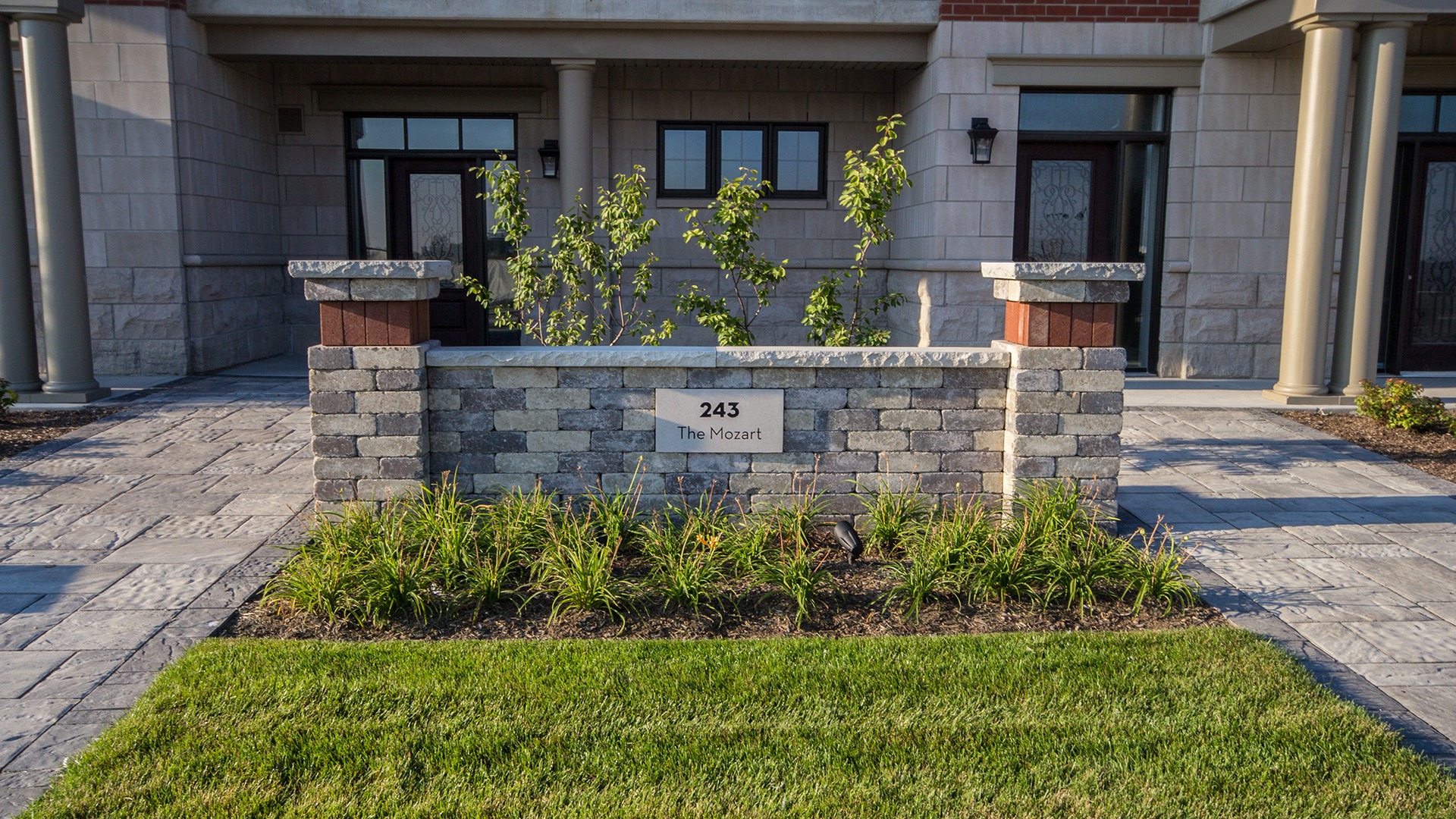 A stone entrance with "243 The Mozart" sign, surrounded by small plants and columns, leads to a building with large windows.