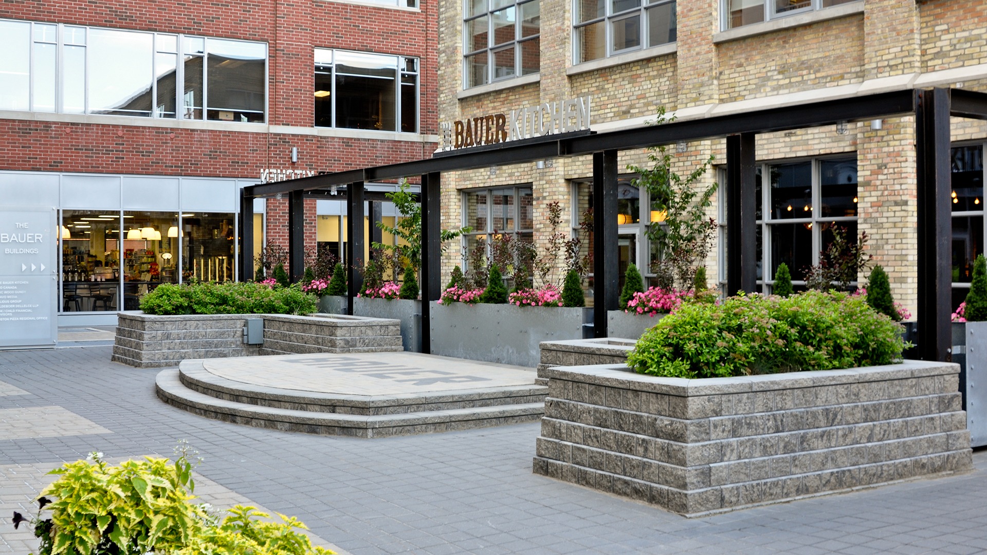 Bauer Buildings' plaza with tiered steps, flower planters, and modern brick architecture. Visible signs include "The Bauer Kitchen."