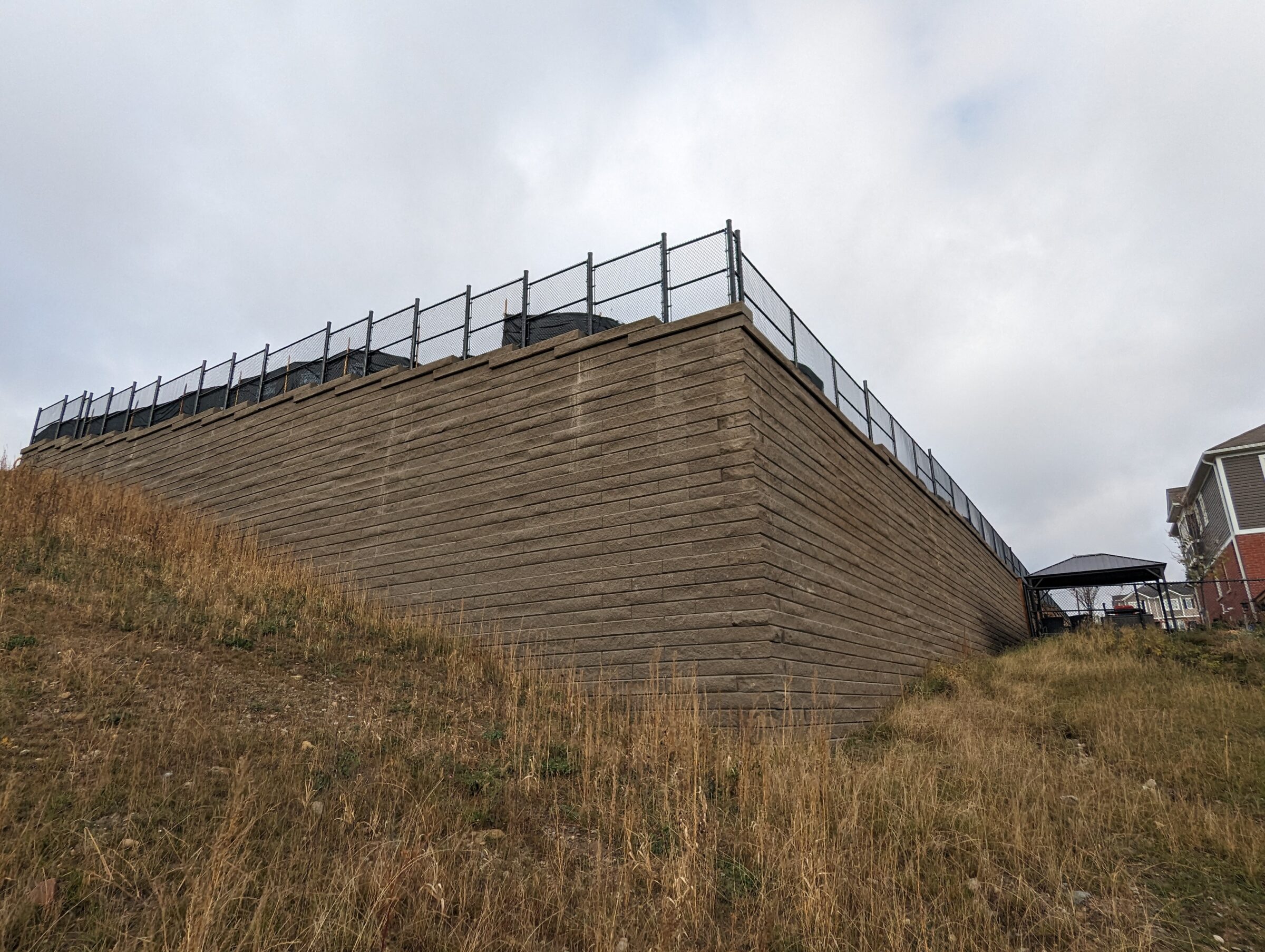 A retaining wall with a chain-link fence overlooks a dry, grassy slope. A person stands under a gazebo structure. Overcast sky above.