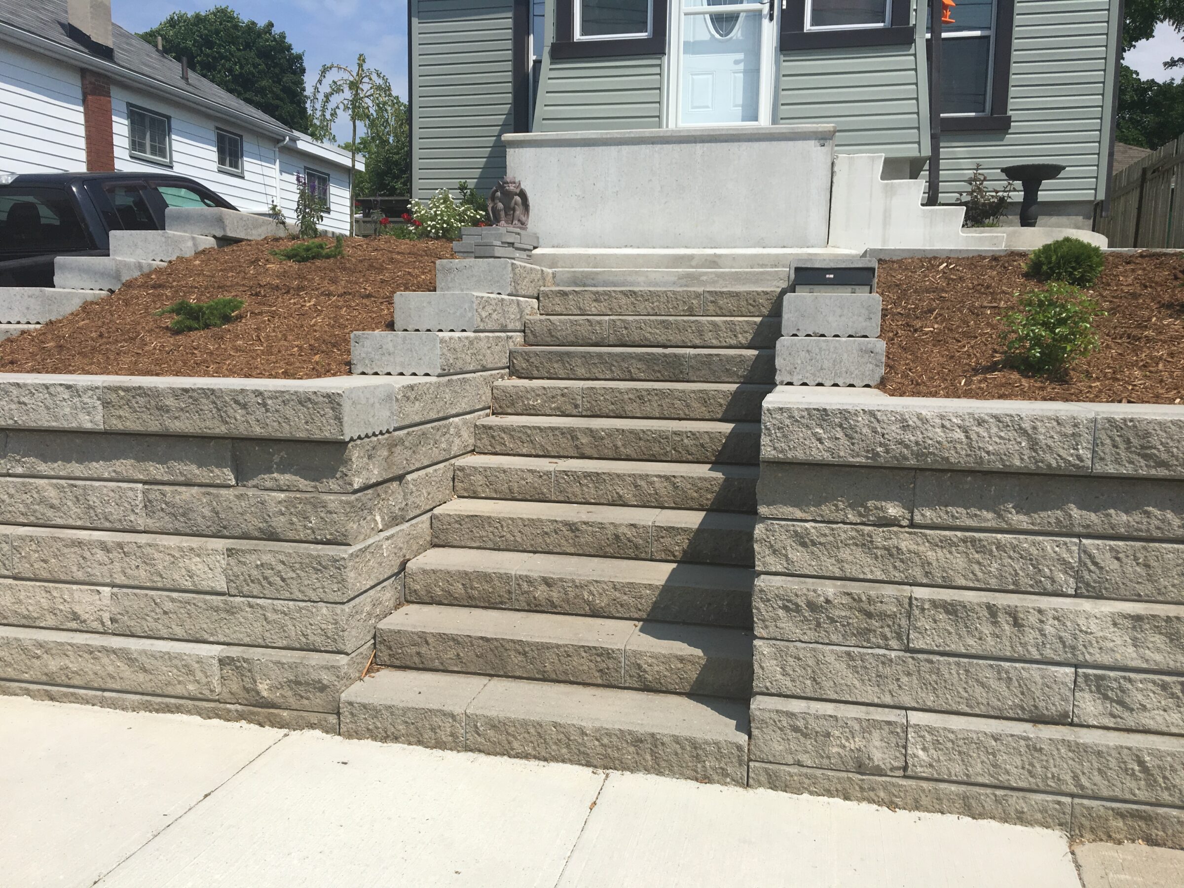 Concrete steps leading to a house entrance, flanked by retaining walls and a garden. A small gargoyle sculpture sits on the left.