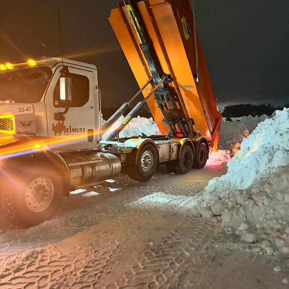 A truck with bright lights is unloading snow in a snowy area at night, surrounded by large piles of snow.